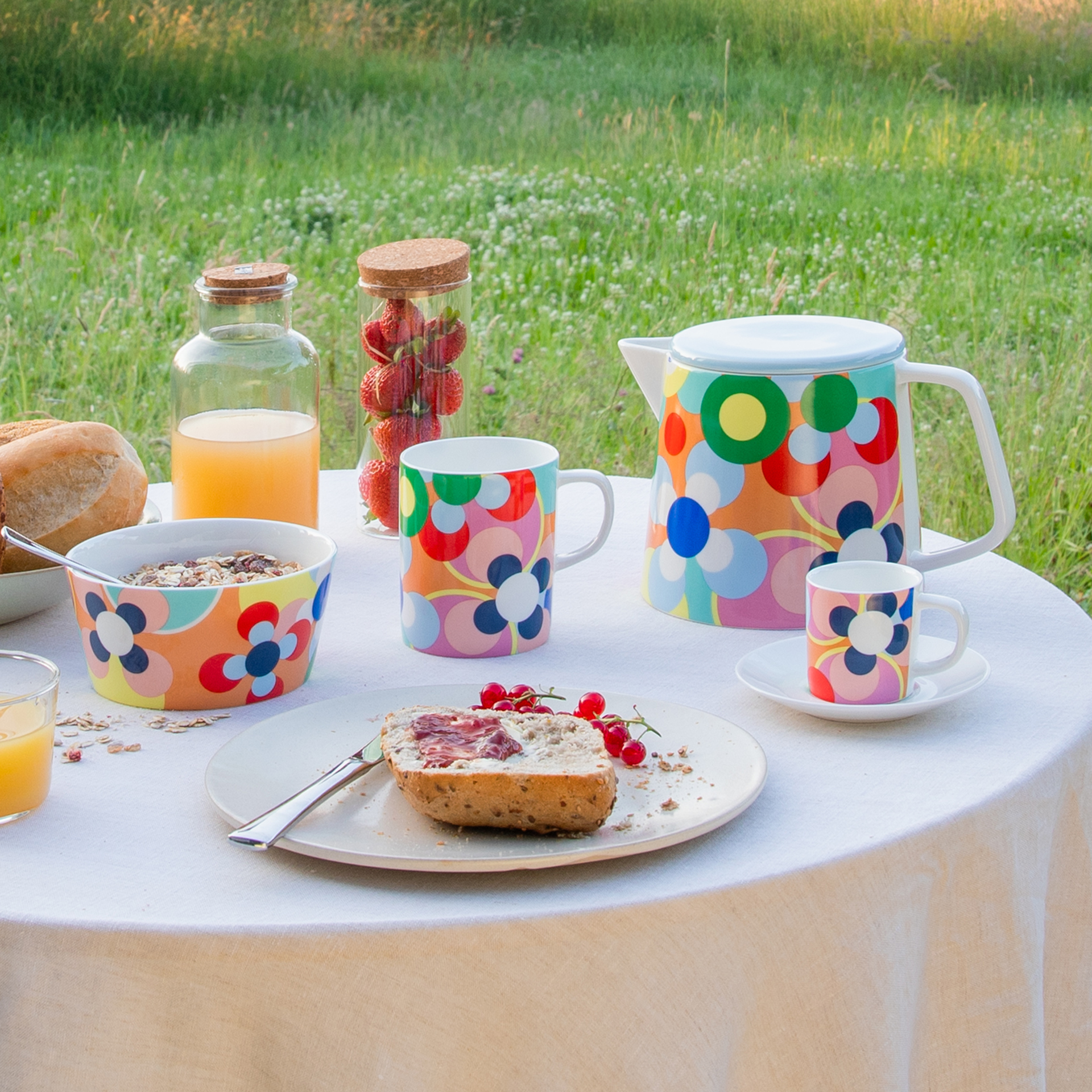 Petit déjeuner au vert : table avec céréales, jus de fruits, fraises, pain avec confiture et vaisselle colorée avec des motifs floraux. Pot, tasses et bol sont à disposition.