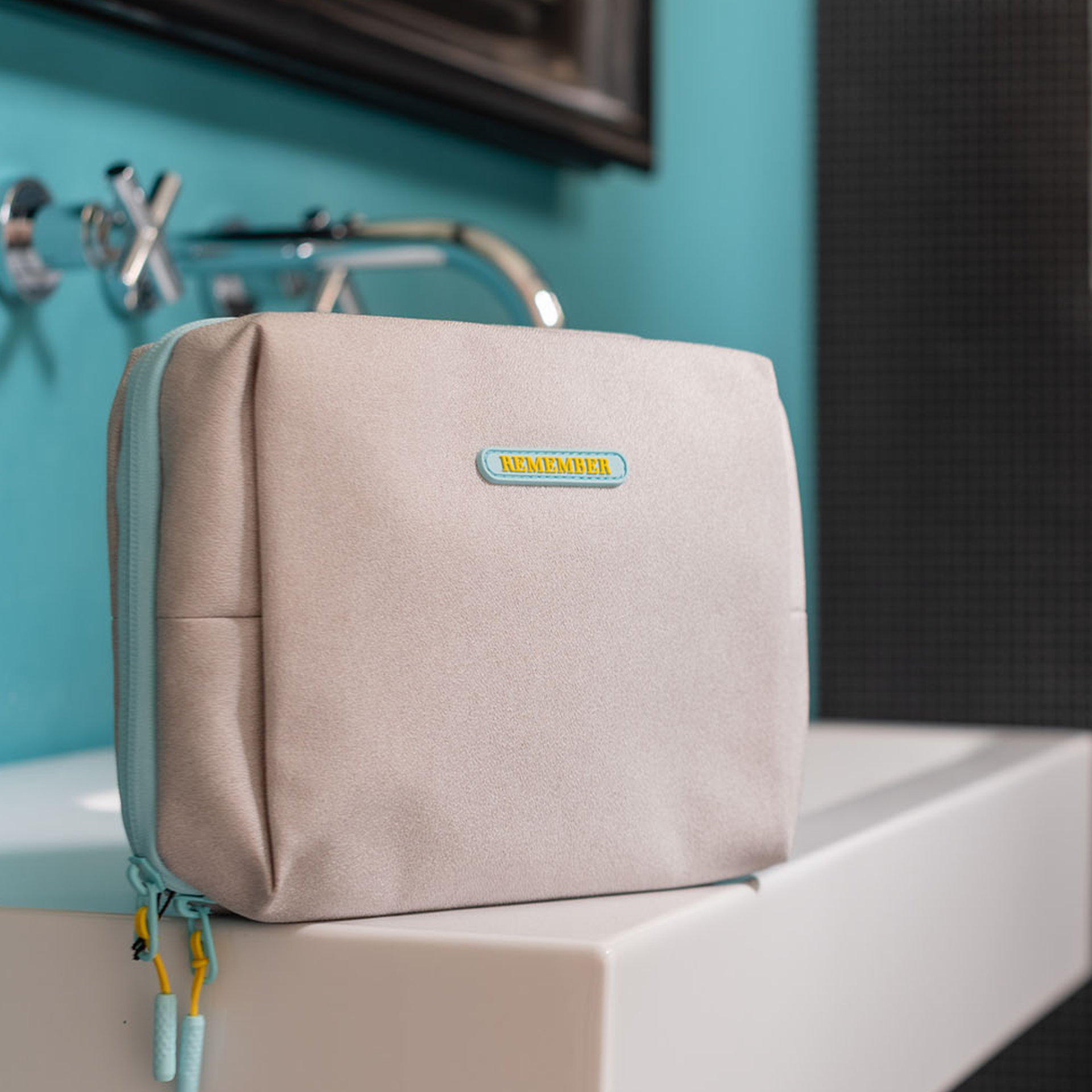 Beige Kulturtasche 'Ocean' mit blauem Reißverschluss und 'Remember'-Logo auf einem weißen Waschtisch, im Hintergrund eine blaue Wand mit Armaturen.