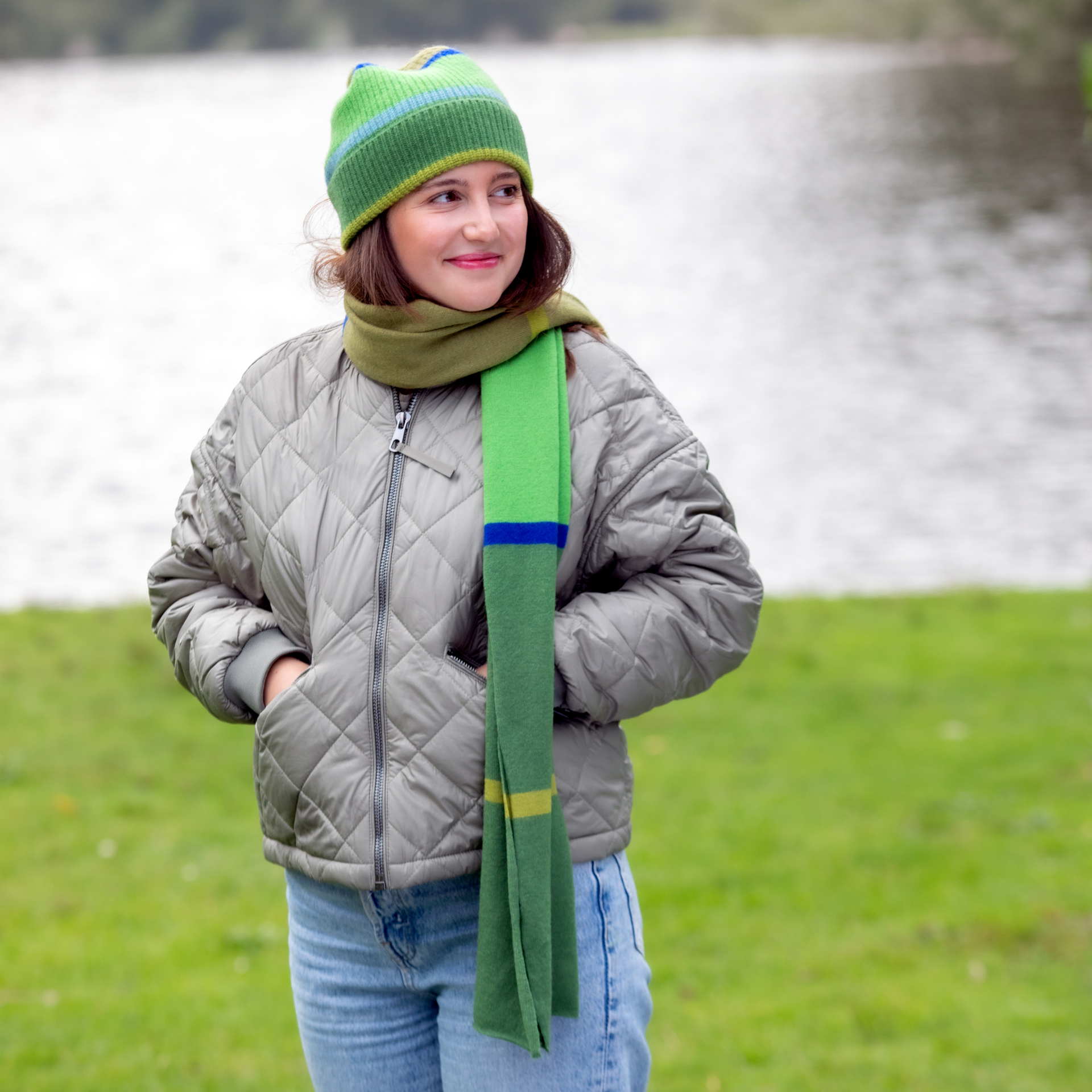 Eine junge Frau mit grüner Mütze und Schal steht auf einer Wiese vor einem See. Sie trägt eine gesteppte Jacke und Jeans und blickt lächelnd nach rechts.