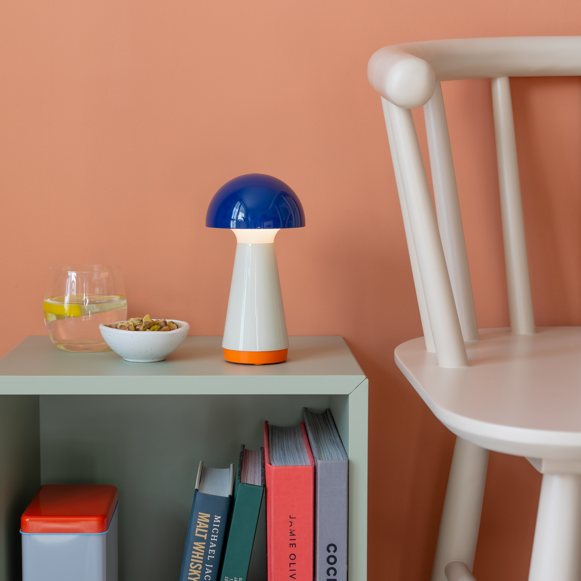 Blaue Tischleuchte 'Bobbi' im Regal mit Büchern, Getränk und Snacks. Die Lampe hat eine pilzförmige Form und spendet Licht. Im Hintergrund ist ein weißer Stuhl vor einer orangefarbenen Wand zu sehen.