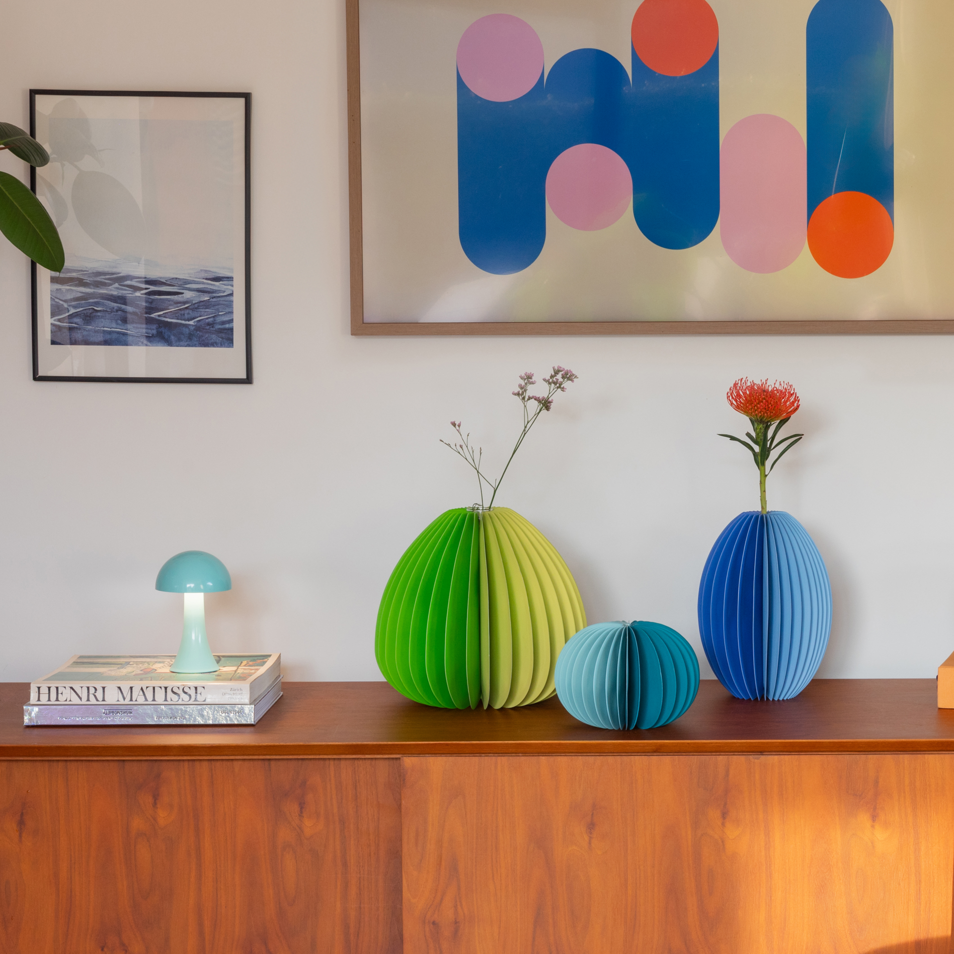 Auf einem Holzschrank sind Vasen in Blau-, Grün- und Türkistönen mit Blumen platziert. An der Wand sind zwei gerahmte Kunstwerke gehängt. Eine blaue Lampe ist auf Büchern positioniert.