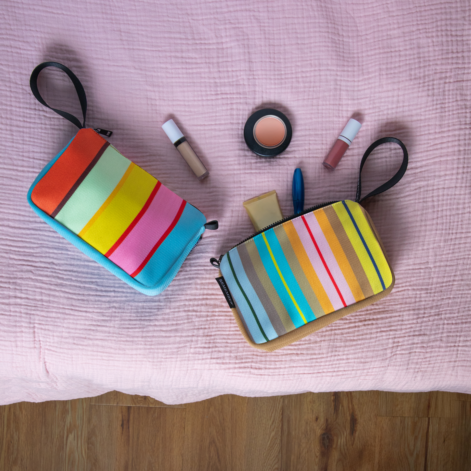 Deux trousses de maquillage Ventara aux couleurs vives, à motif rayé sur tissu rose. L'une a des côtés bleus, l'autre beiges, avec des poignées noires. Entre elles : rouge à lèvres, mascara, correcteur, poudre. L'une est ouverte.