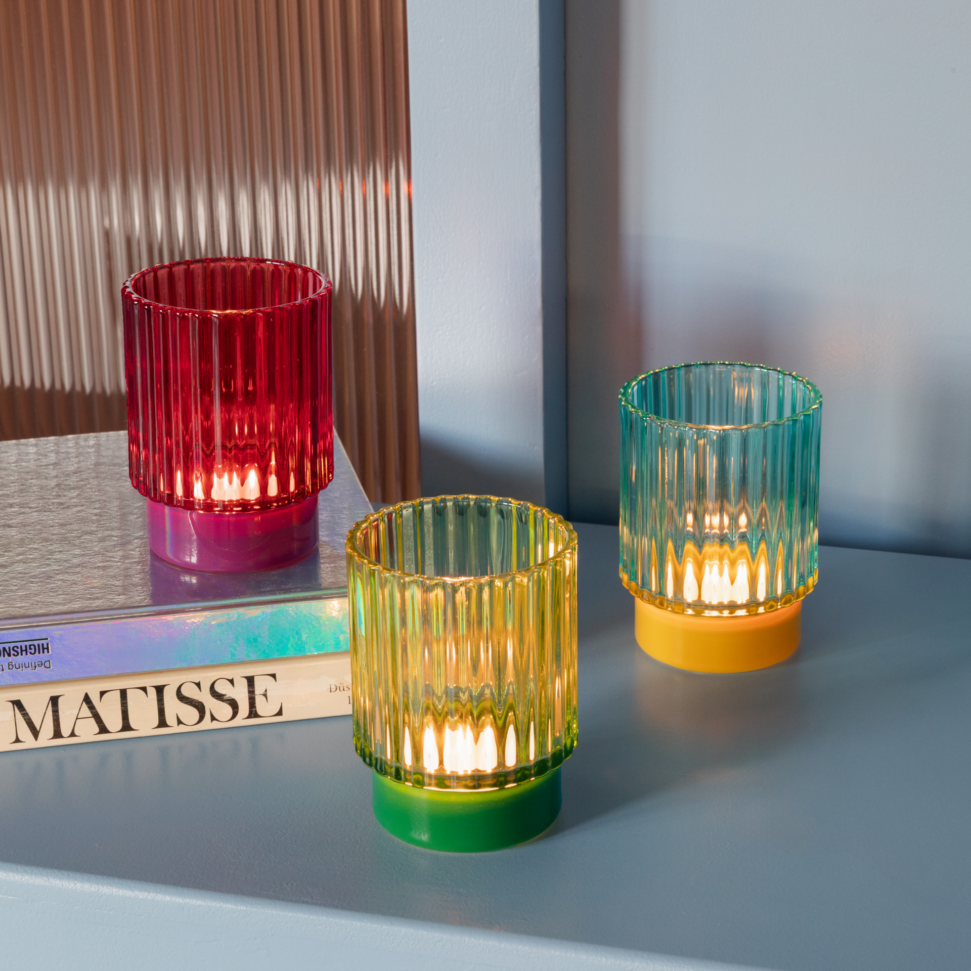 Drei farbige, gerippte Glaswindlichter mit farblich passenden Sockeln stehen auf einem Tisch. Sie sind rot, gelb und blaugrün und scheinen von innen beleuchtet.