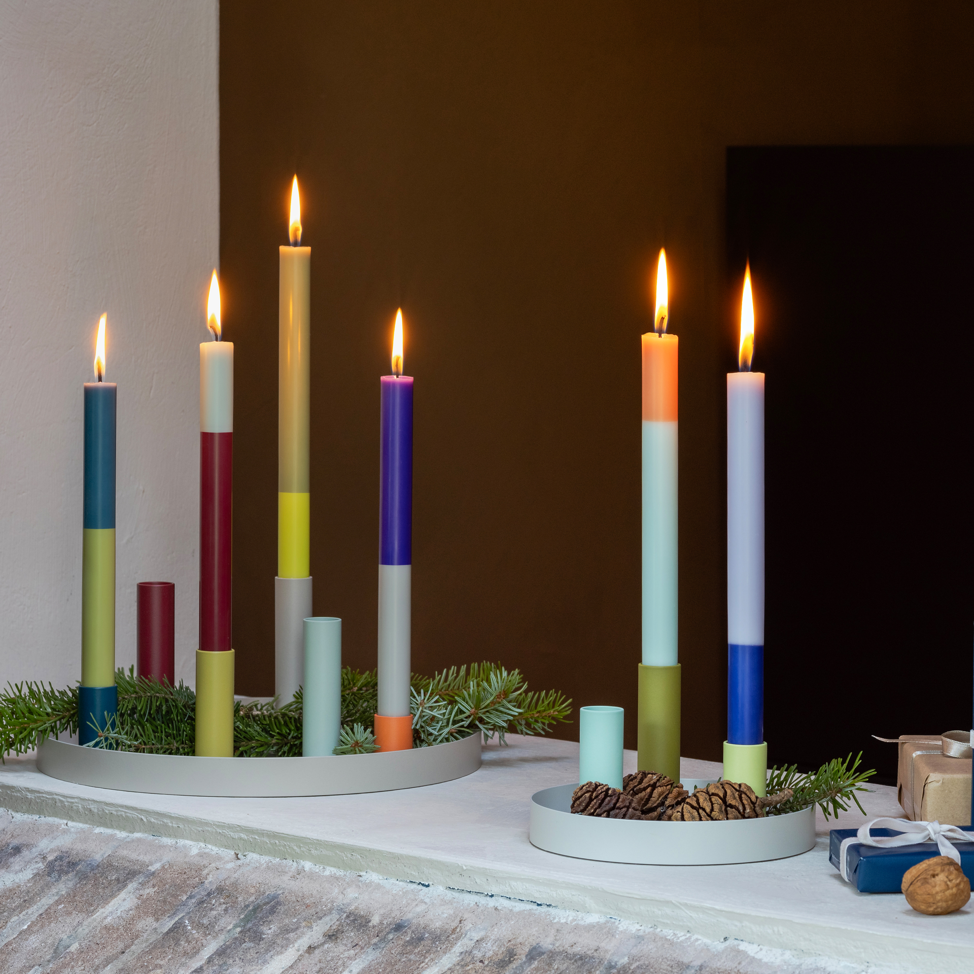 Brennende Stabkerzen 'Oslo' in farbenfrohem Color-Blocking-Design, arrangiert auf runden Tabletts mit Tannenzweigen. Einige Kerzen sind unterschiedlich hoch und manche brennen.