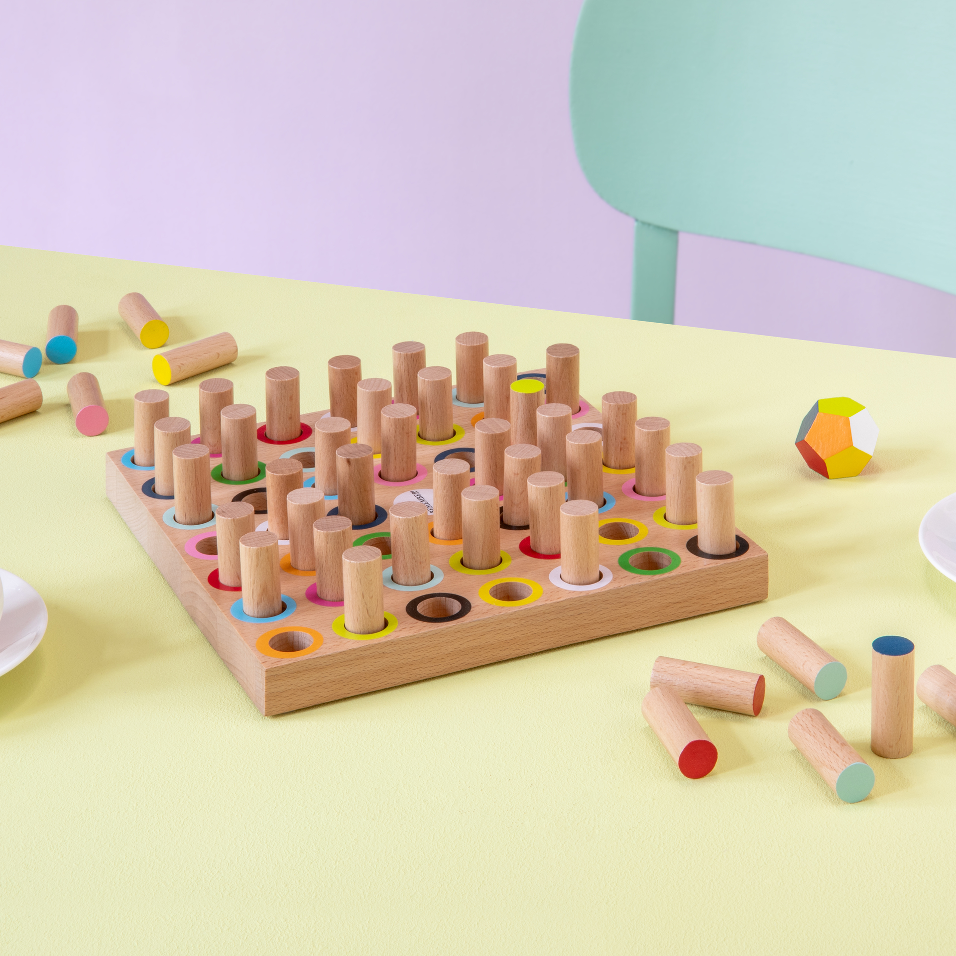 Das Produkt zeigt Holzspielzeug Momo mit farbigen Kreisen und Holzstiften auf einem hellgelben Tisch. Einige Stifte liegen daneben. Eine bunte Kugel und ein mintgrüner Stuhl sind im Hintergrund zu sehen.
