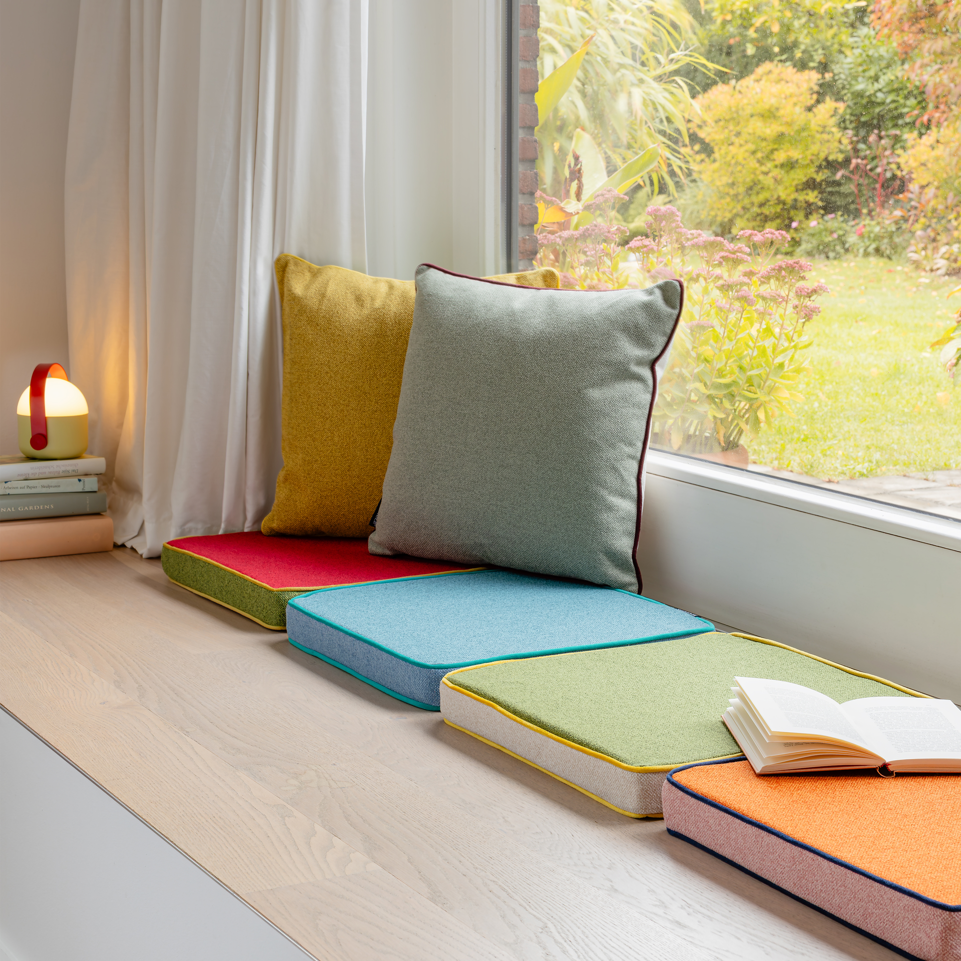 Fensterbank mit bunten Kissen und zwei Kissen am Fenster. Ein aufgeschlagenes Buch liegt auf einem Kissen. Draußen ist ein Garten zu sehen.