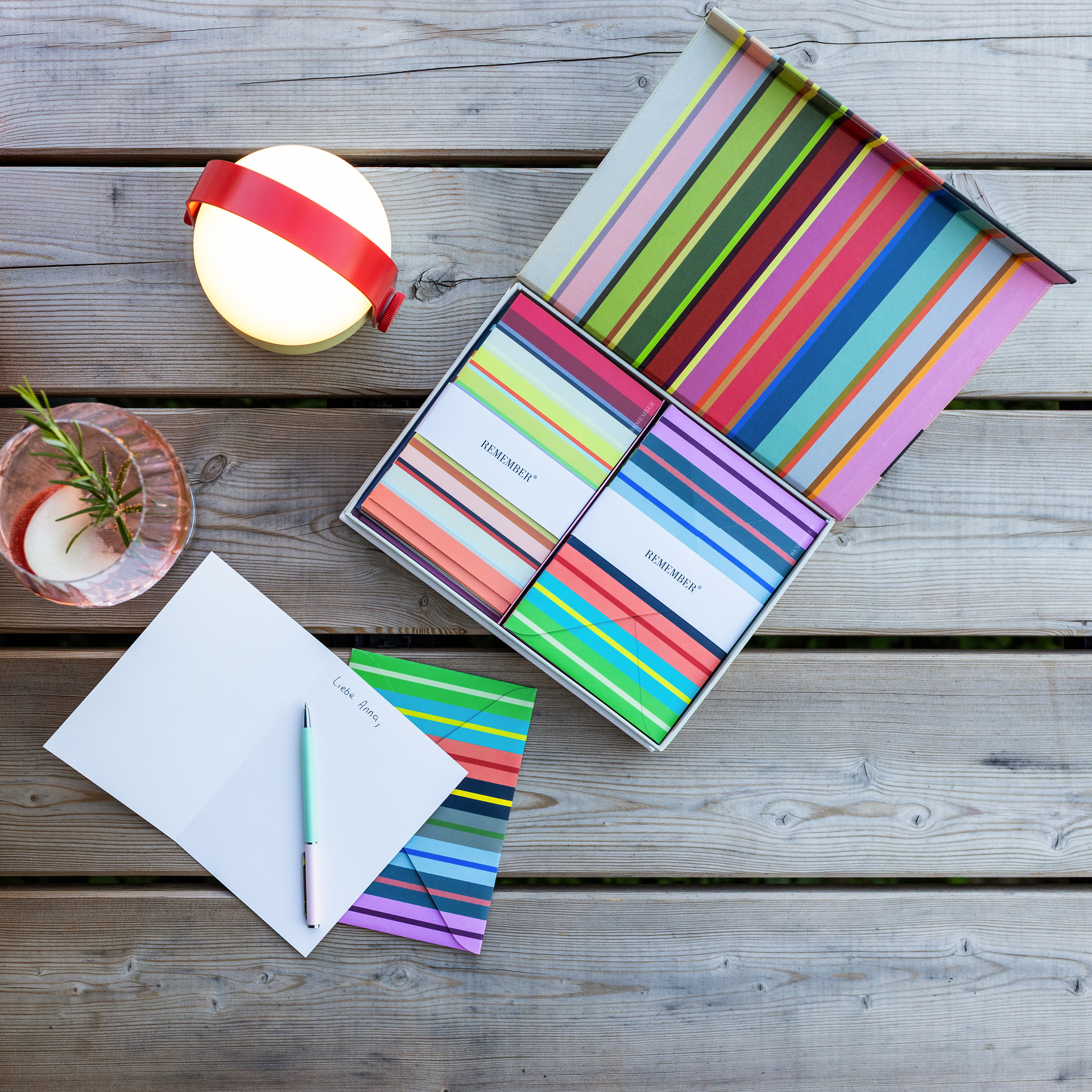 Boîte magnétique ouverte avec piles de cartes pliables rayées et enveloppes assorties. Chaque pile est entourée d’un ruban blanc «REMEMBER ». À droite, une carte dépliée au même motif à rayures.