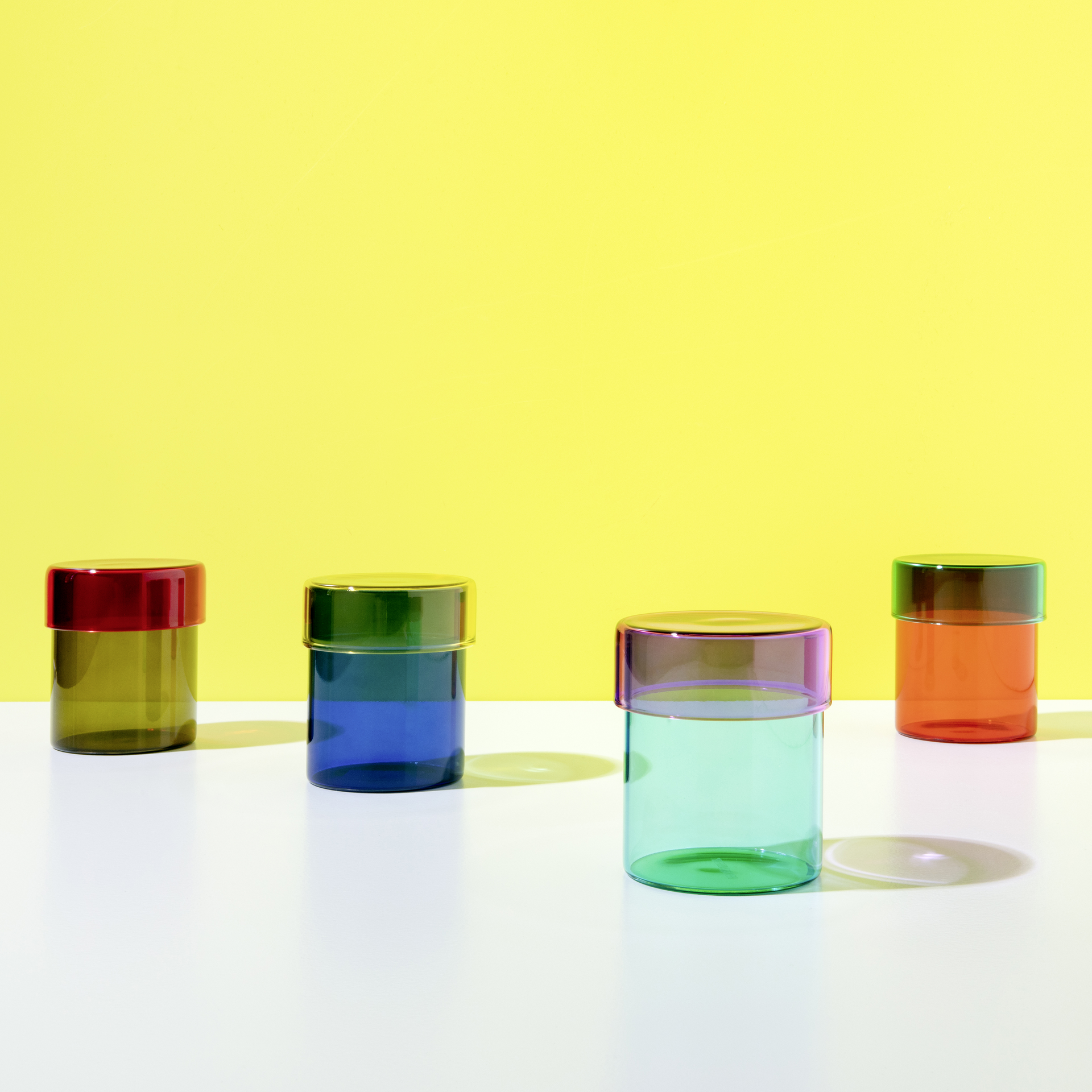 Vier farbige, zylindrische Glasdosen mit Deckel stehen auf einer weißen Oberfläche vor einem gelben Hintergrund.