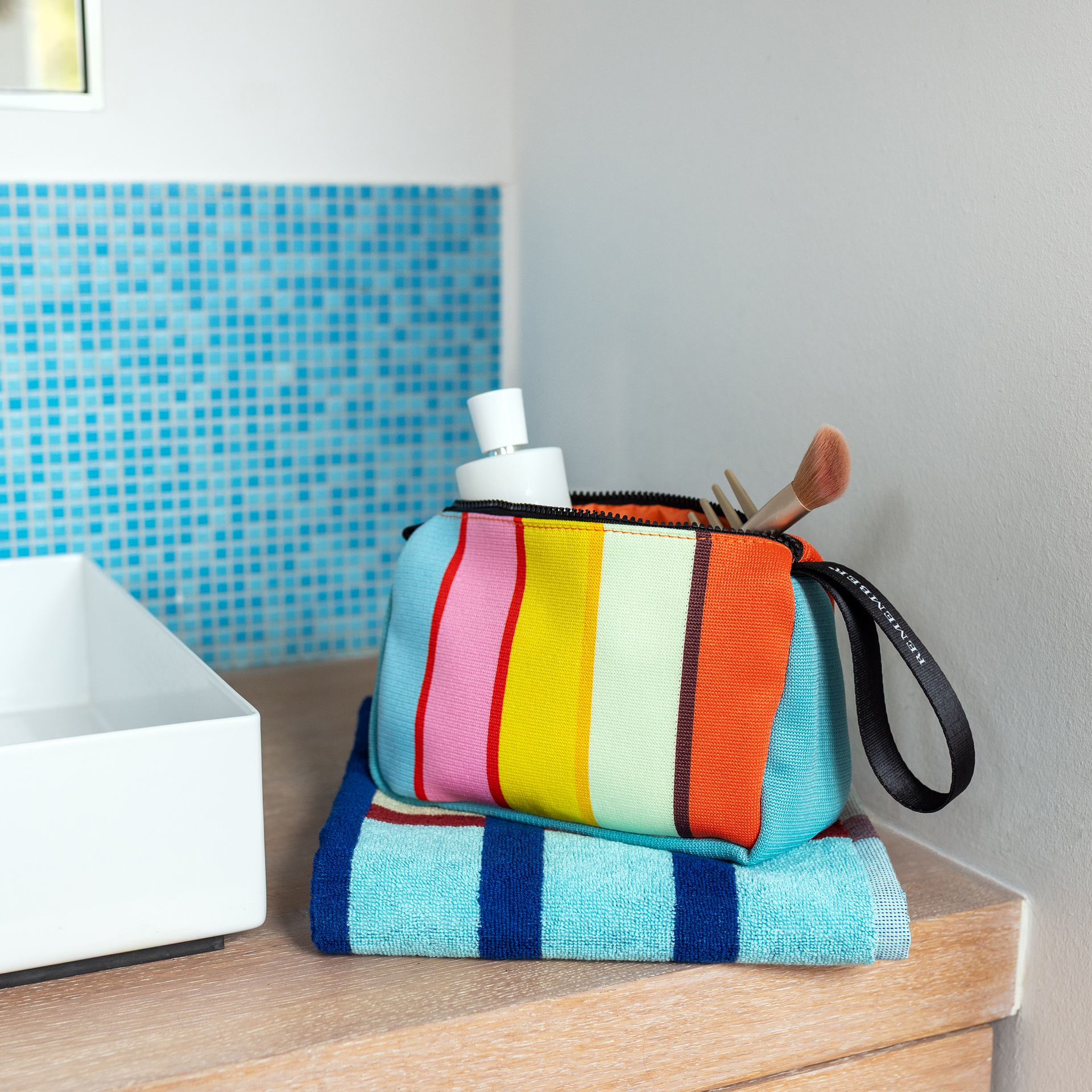 Trousse de maquillage rectangulaire Levante au motif rayé joyeux. Rayures verticales orange, vert clair, jaune et rose, rehaussées de fines lignes rouges et marron. Côtés et fond bleu clair. Fermeture à glissière noire et poignée de transport.