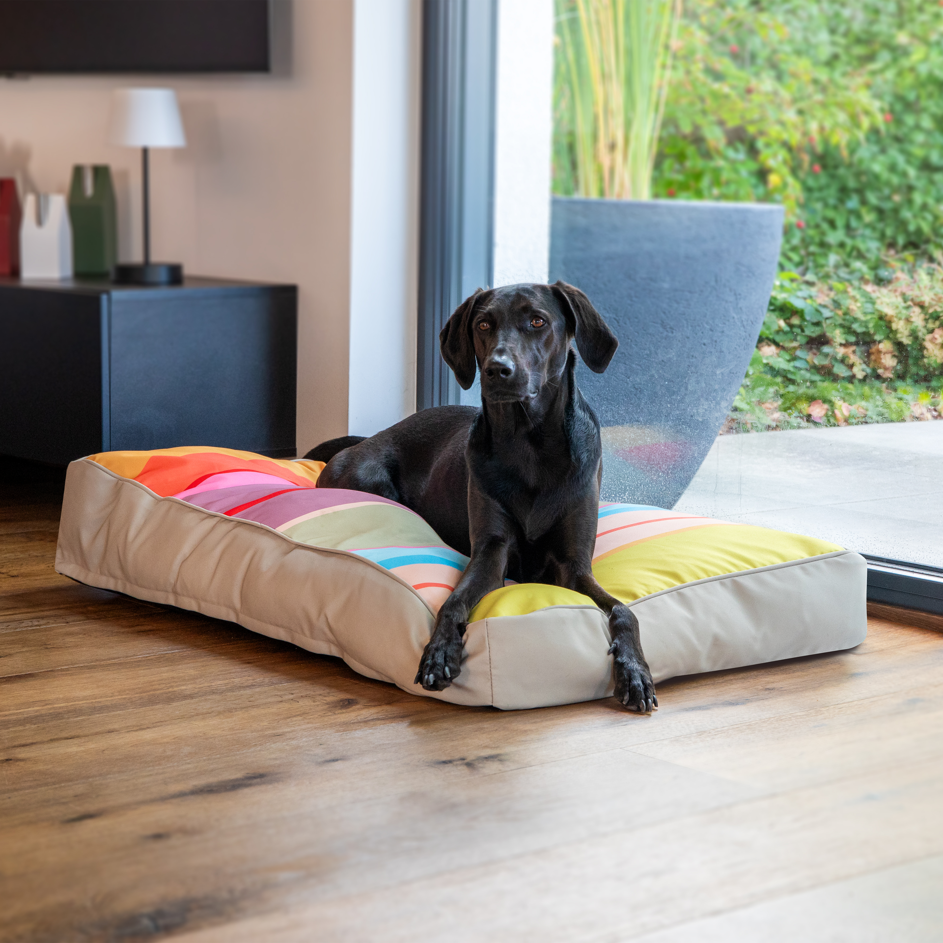 Coussin rectangulaire pour chien Theo, taille L, base robuste beige et dessus coloré accrocheur. De larges rayures horizontales jaunes, orange, rouges, roses, violettes, vertes et bleu clair offrent un repos tranquille.