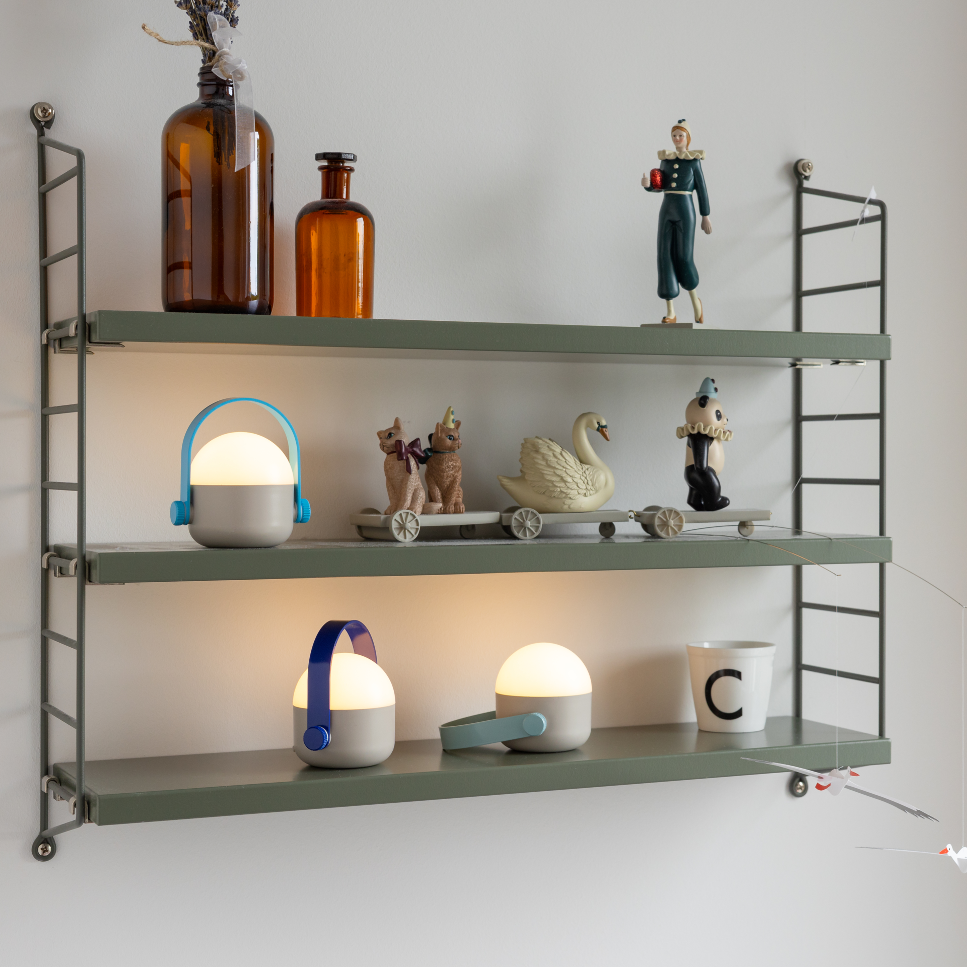 Drei leuchtende OLE mini blue LED-Leuchten im 3er-Set stehen auf einem olivgrünen Regal vor weißer Wand. Akkubetrieben, mit hellgrauem Sockel, milchweißer Kugel und Bügeln in Hellblau, Dunkelblau und Mintgrün. Warmweißes Licht.