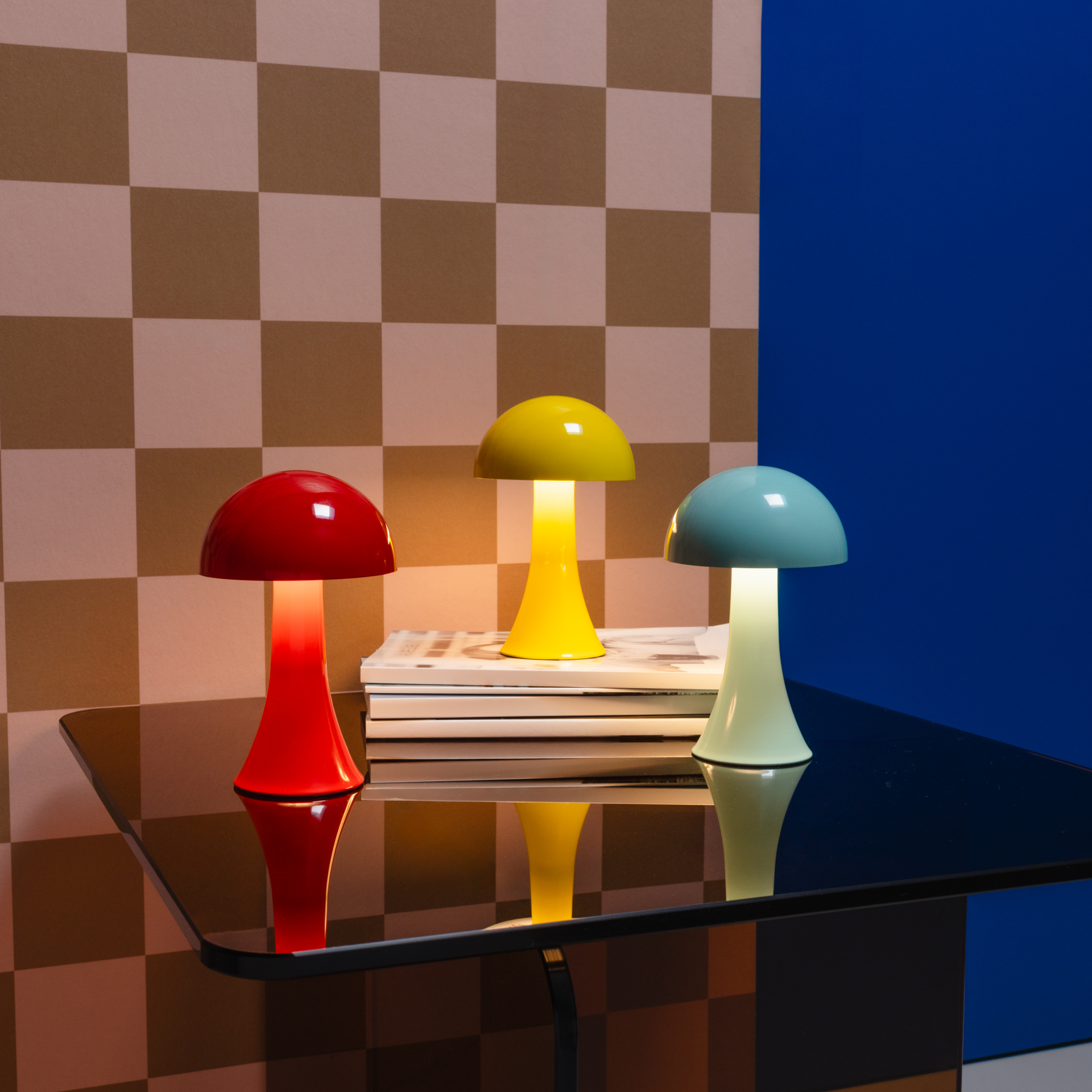 Drei pilzförmige Leuchten in Rot, Gelb und Blau sind auf einer glänzenden Tischplatte platziert. Im Hintergrund befindet sich eine Wand mit Schachbrettmuster sowie eine blaue Fläche.