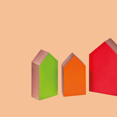 Drei farbige Holzhaus-Figuren (grün, orange, rot) stehen auf einer einfarbig hellbraunen Fläche. Die Häuser sind unterschiedlich groß und haben braune Dächer.