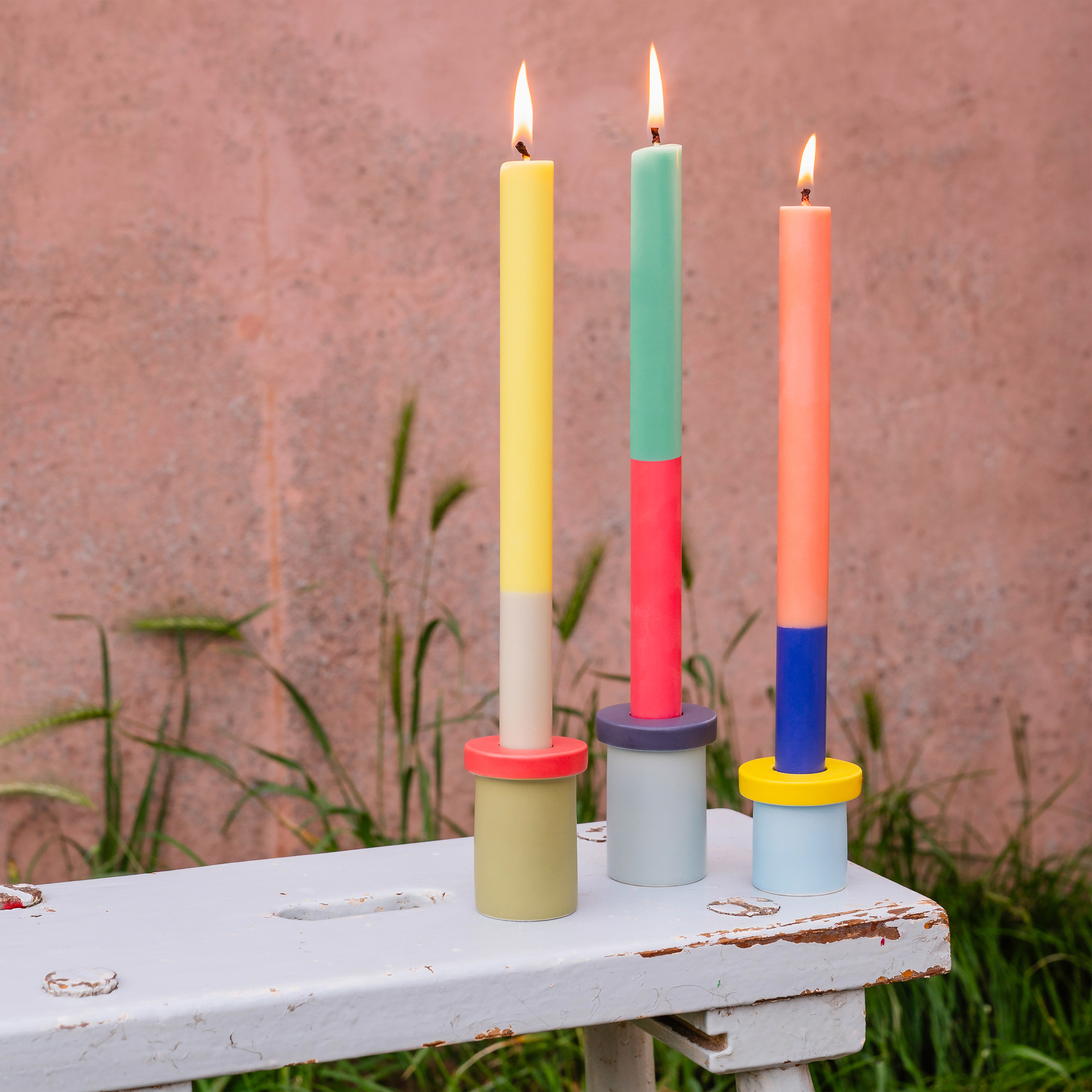 Drei bunte, brennende Stabkerzen im Color-Blocking-Design, stehend auf Kerzenhaltern mit runden Elementen, auf einem weiß gestrichenen Tisch vor rosafarbenem Hintergrund.