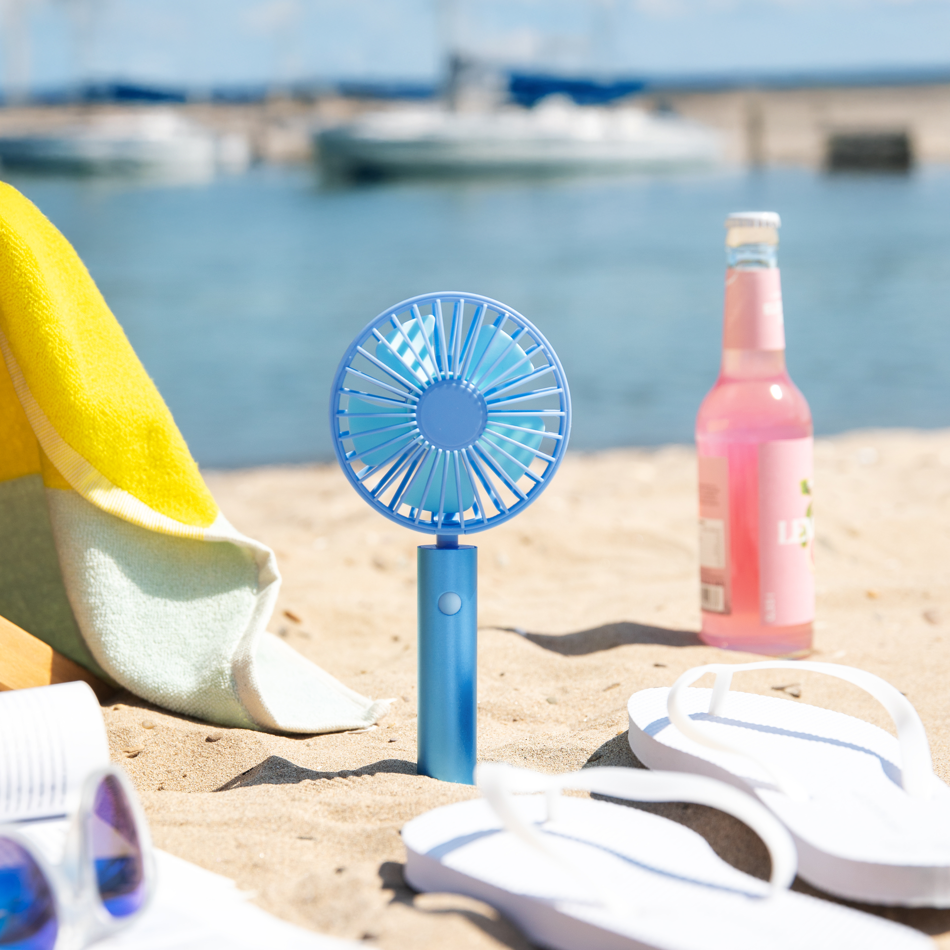 Un ventilatore blu a batteria ricaricabile "Flow aqua" si trova sulla spiaggia. Sullo sfondo si vedono il mare, le barche e un asciugamano. In primo piano, infradito e una bibita.