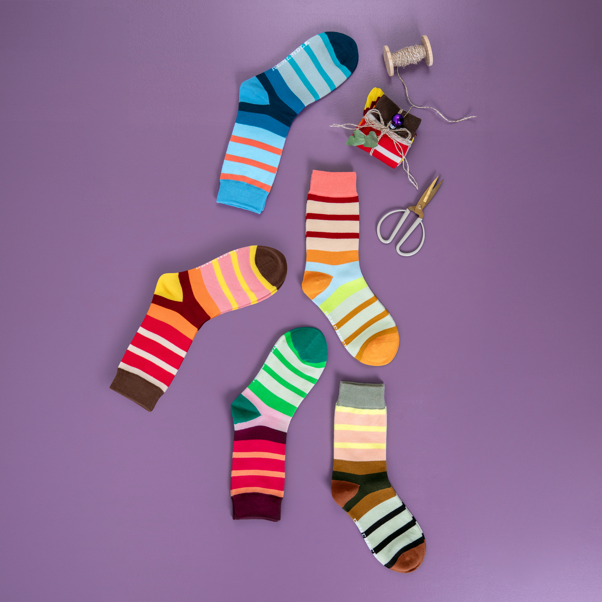 Draufsicht: Mehrere bunte Socken Modell 70 in verschiedenen Designs auf lila Hintergrund, daneben ein Geschenk, Fadenrolle und Schere.