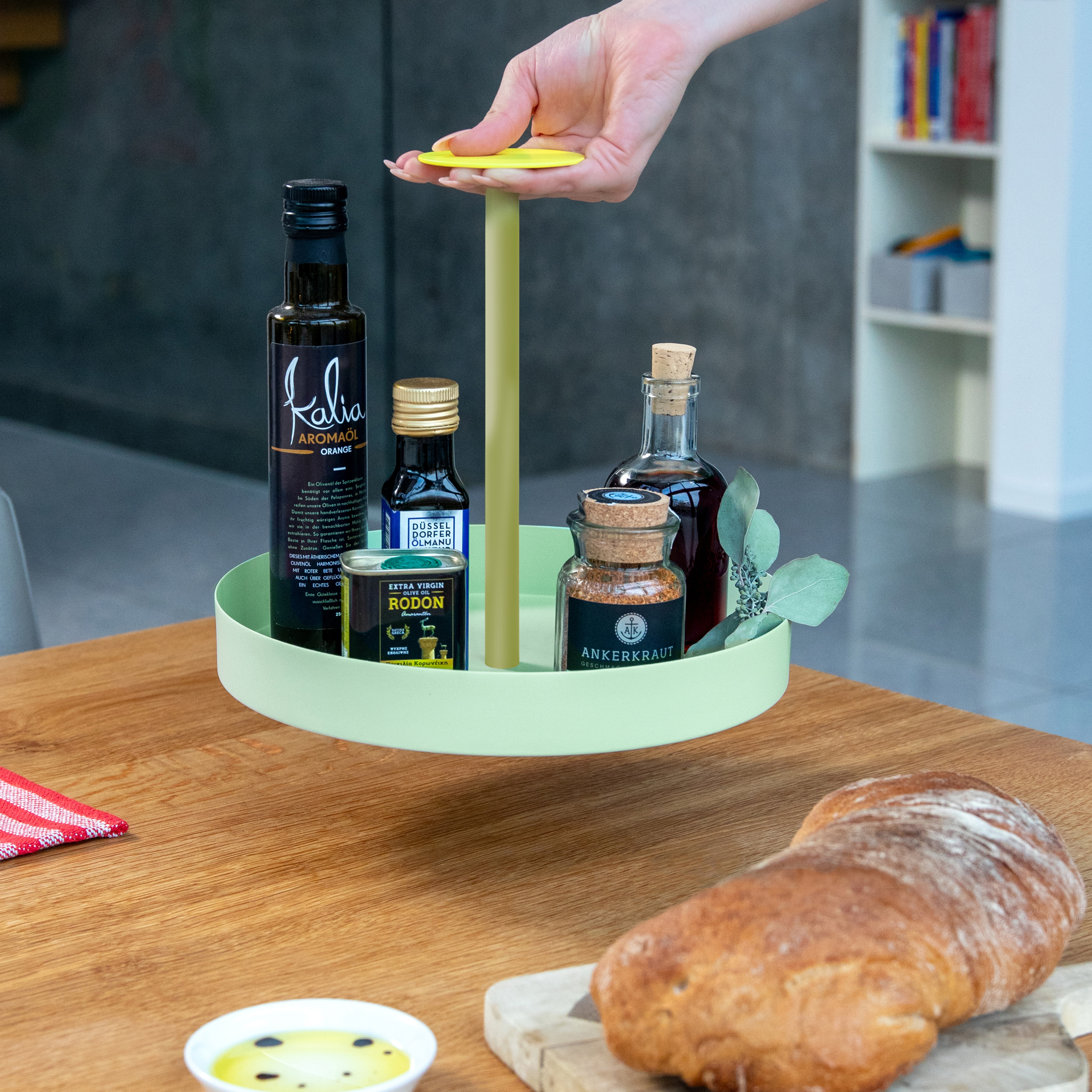 Das limefarbene Tablett 'Nana' mit Griff, Ölflaschen und Gewürzen auf einem Holztisch, Brot und Dip im Vordergrund. Eine Hand greift nach dem Griff des Tabletts.