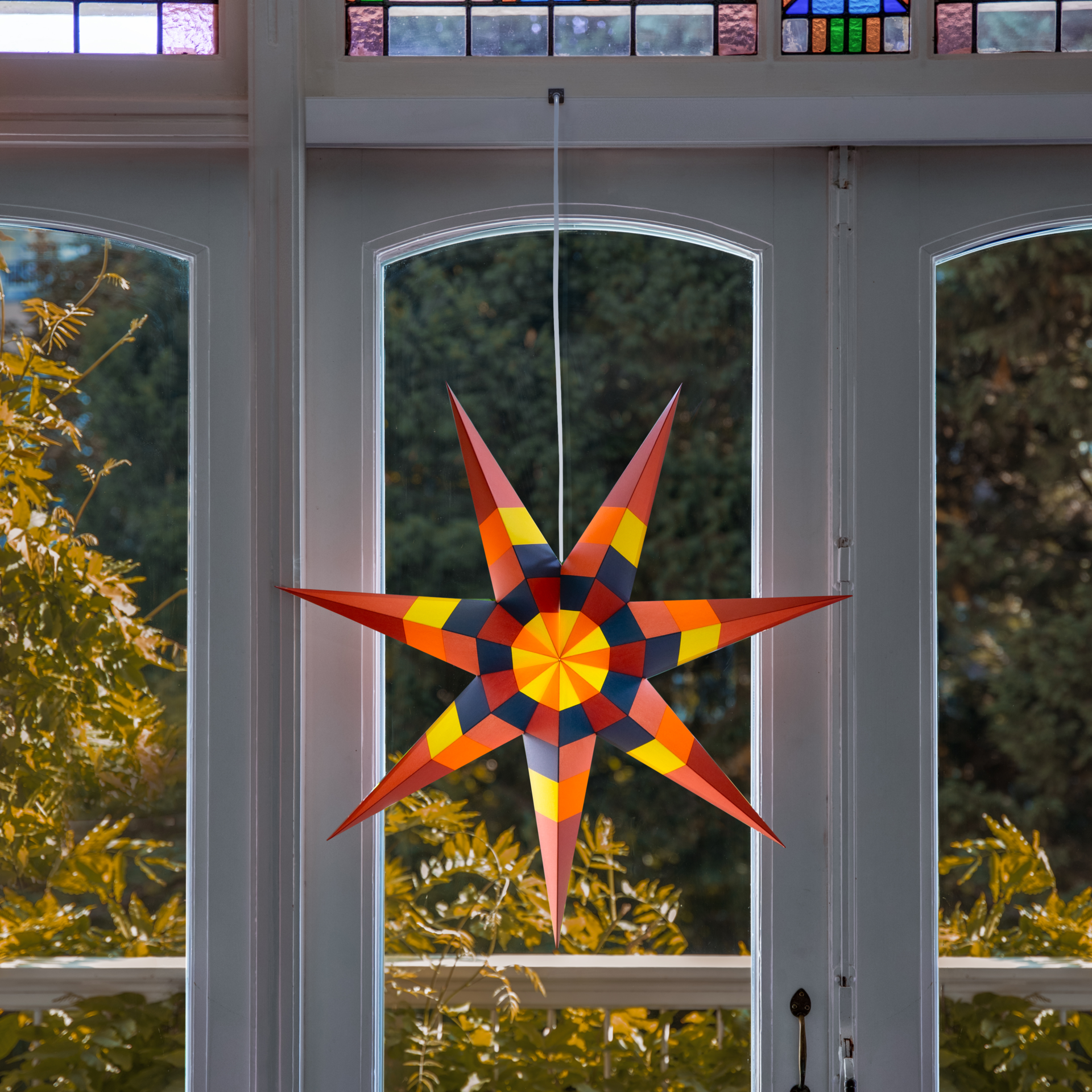Farbenfroher Papierstern mit sieben zackigen Spitzen, Oberfläche aus vielen gefalteten geometrischen Flächen in Rot, Orange, Gelb, Dunkelblau und Rosa. Radiales Muster um die Mitte. Oben weiße Kordel, unten silberner Stopper.