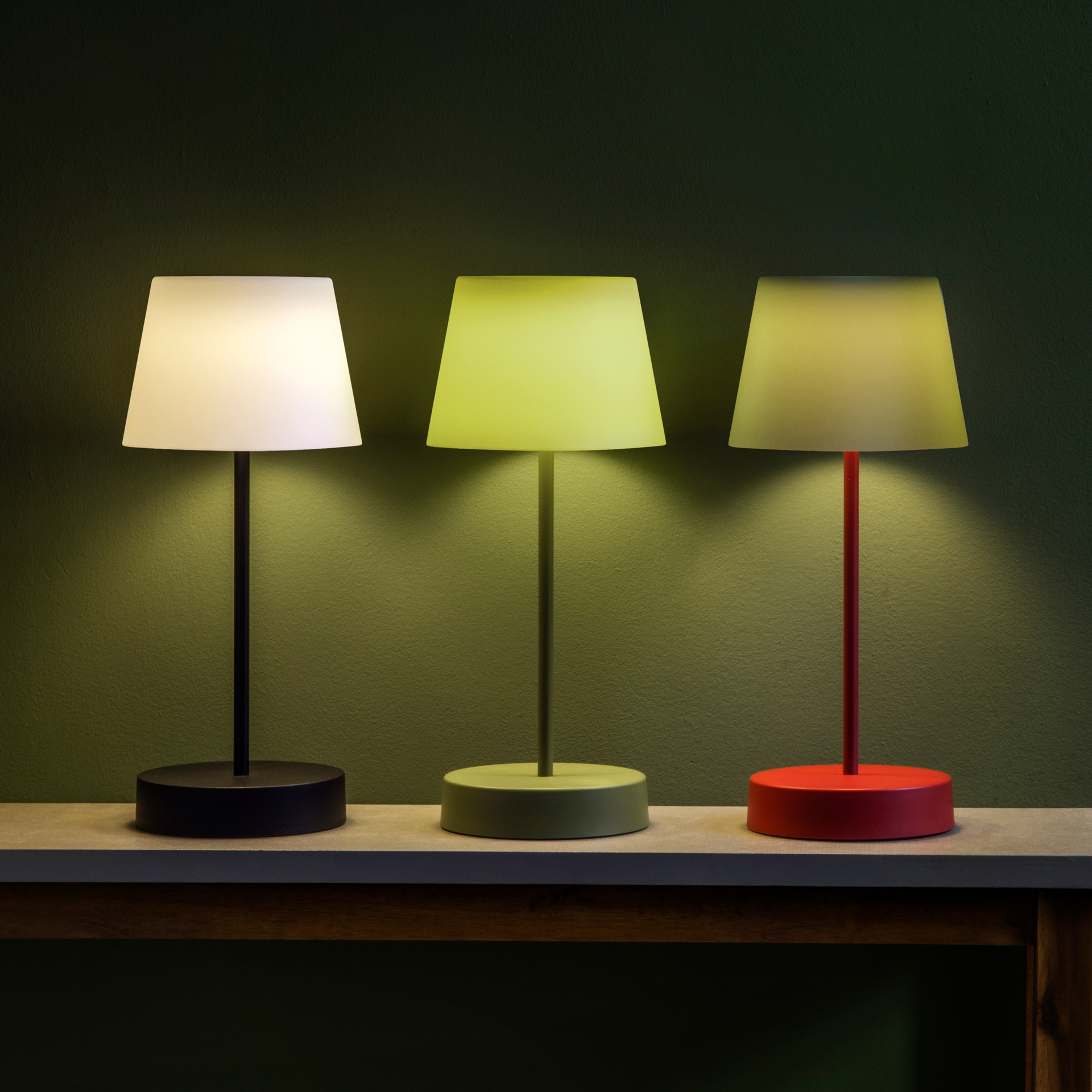 Drei 'Oscar' Tischleuchten in Weiß, Grün und Rot leuchten auf einem Tisch. Die Lampen haben einen runden Fuß und einen konischen Schirm und geben warmes Licht ab.