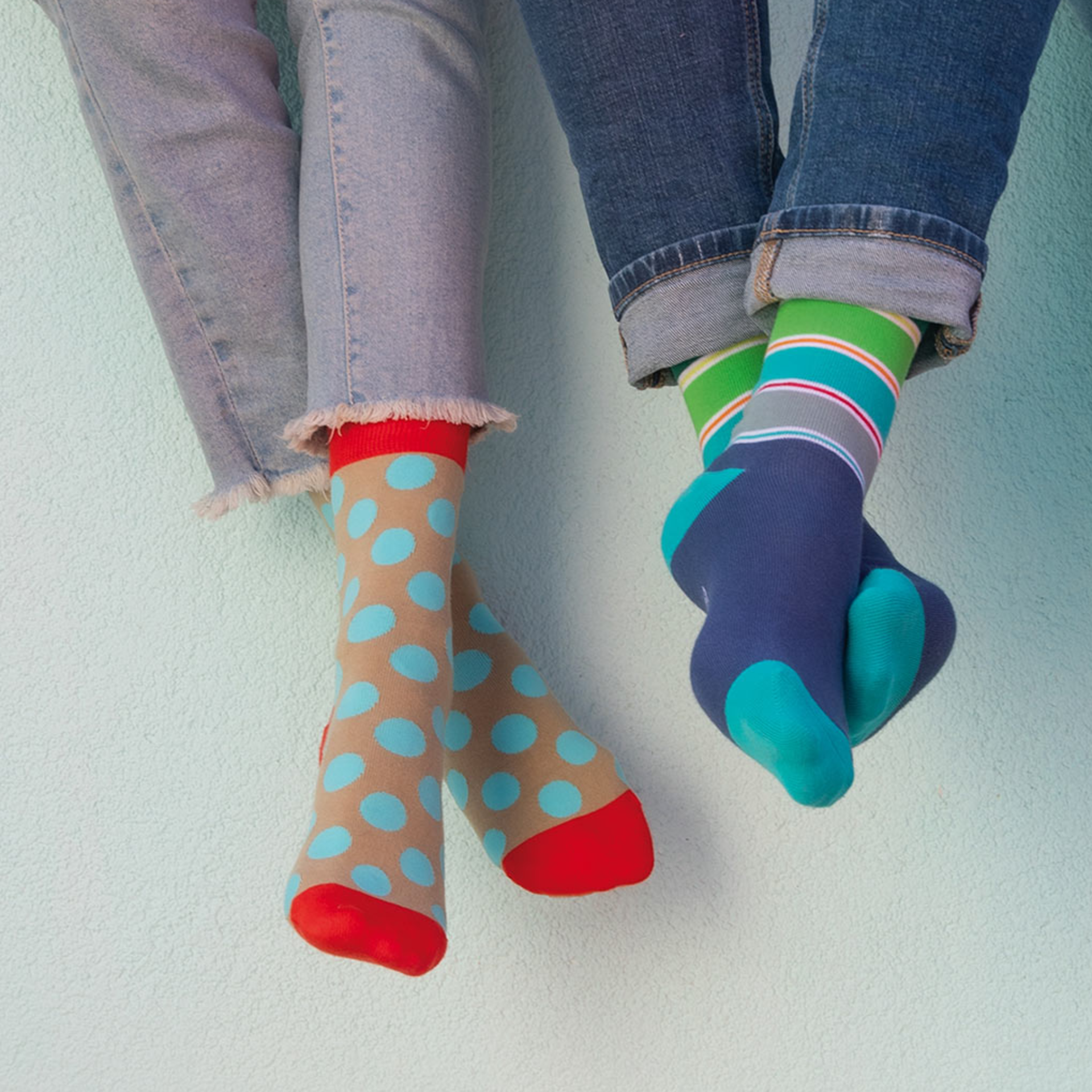 Zwei Paar Füße mit farbenfrohen Socken. Ein Paar mit hellbraunen Socken und blauen Punkten. Das andere Paar mit gestreiften Socken in Grün, Türkis und Grau.