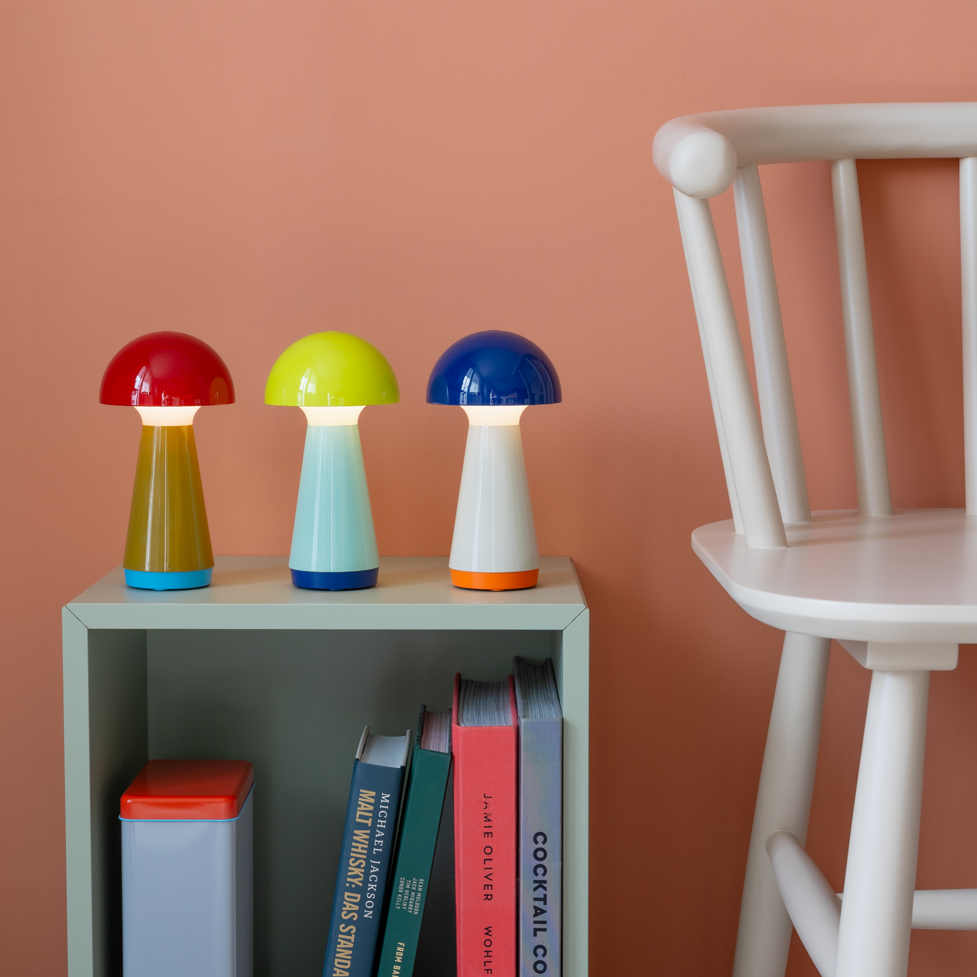 Drei Bobbi Tischleuchten in Rot, Gelb und Blau auf einem Regal, daneben ein Stuhl vor einem orangen Hintergrund. Die Leuchten sind eingeschaltet.