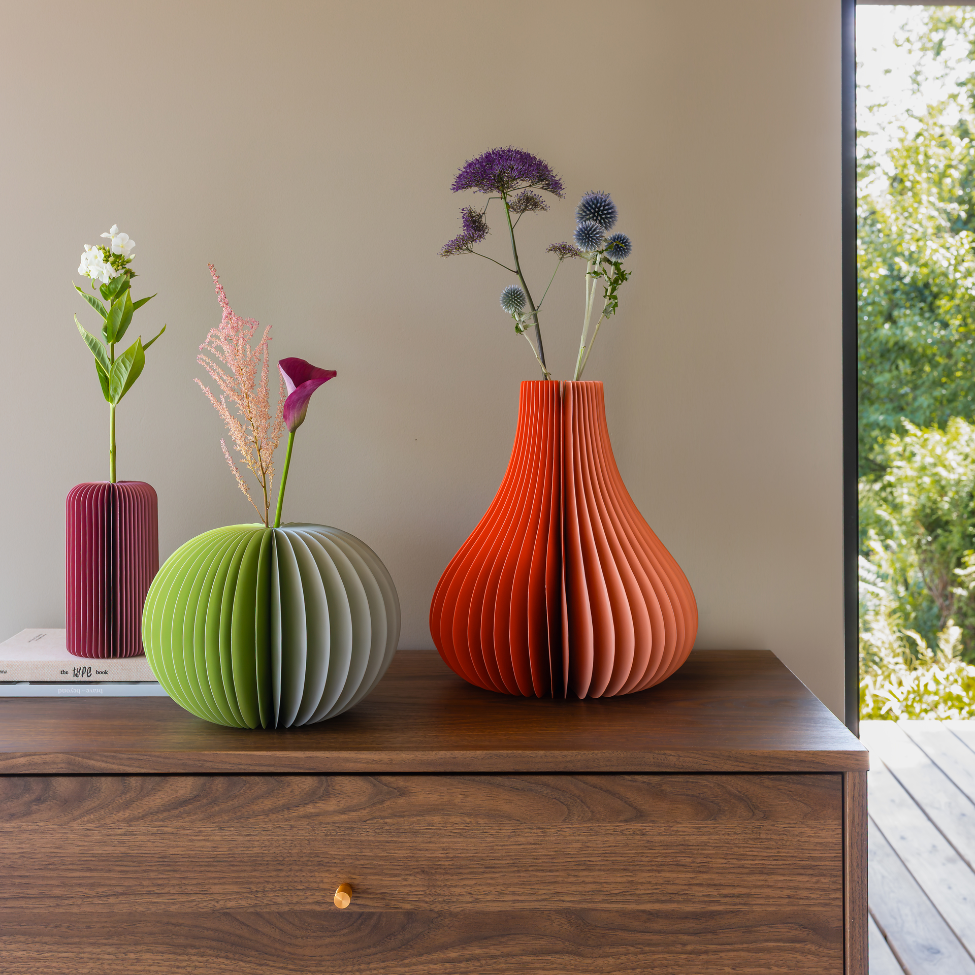 Drei gefaltete Papiervasen auf dunklem Schrank: links bordeaux mit weißen Blüten, mittig hellgrün mit pinker Calla, rechts große orange „Yuki“ mit lila Disteln. Heller Raum, Fenster, Bäume.