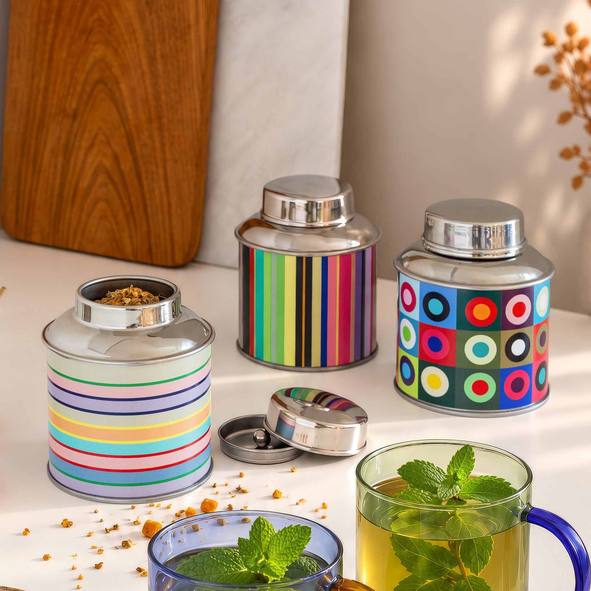 Scatola da tè cilindrica in acciaio inossidabile con coperchio argentato lucido. Il corpo è decorato con un vivace motivo a strisce verticali nei brillanti toni autunnali del verde, giallo, arancione, rosso, blu e grigio.