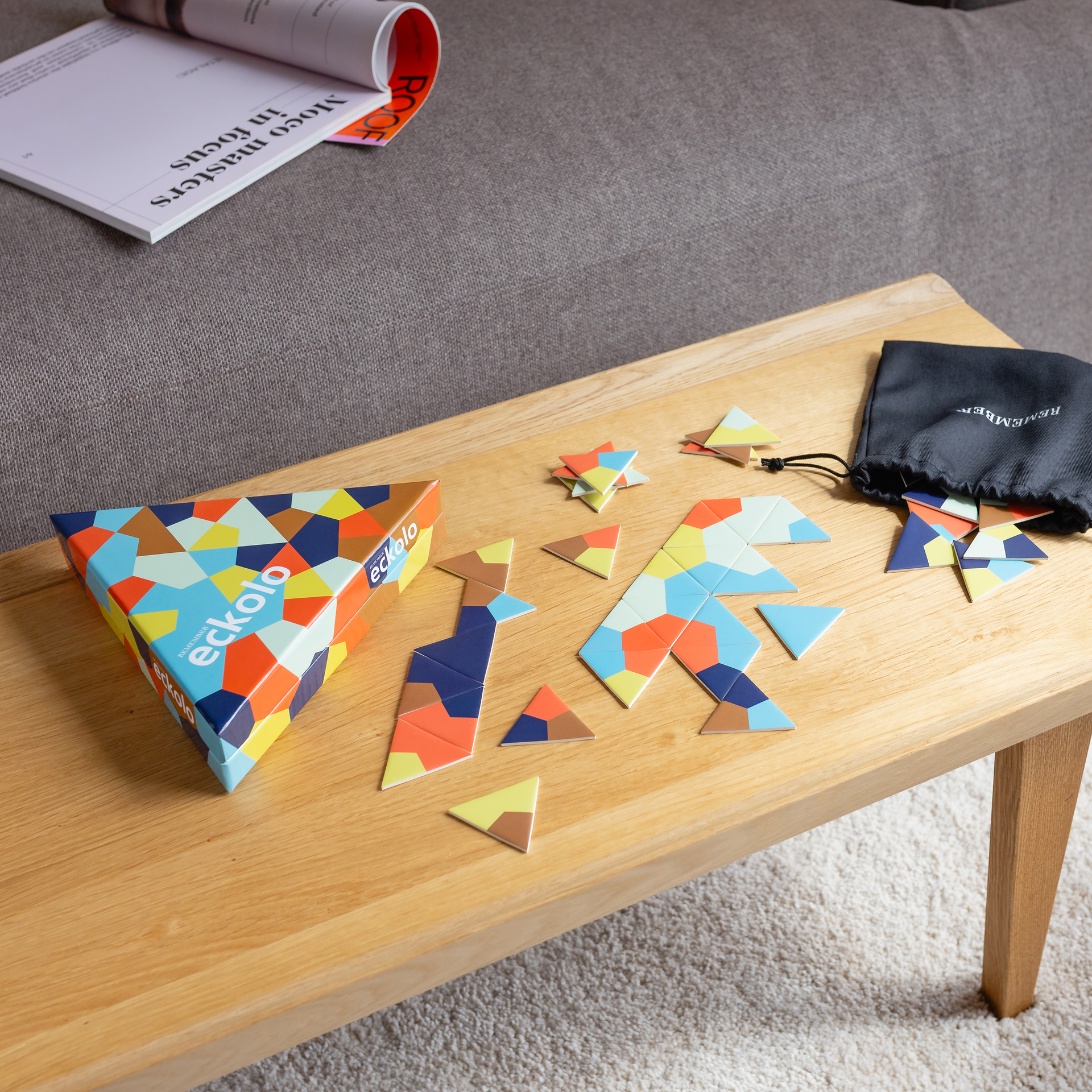 Dreieckige Schachtel des Spiels Eckolo mit buntem geometrischen Muster aus Polygonen in Blau, Orange, Gelb und Braun. Der Schriftzug „eckolo“ ist zweimal sichtbar. Leicht schräg von oben vor weißem Hintergrund.