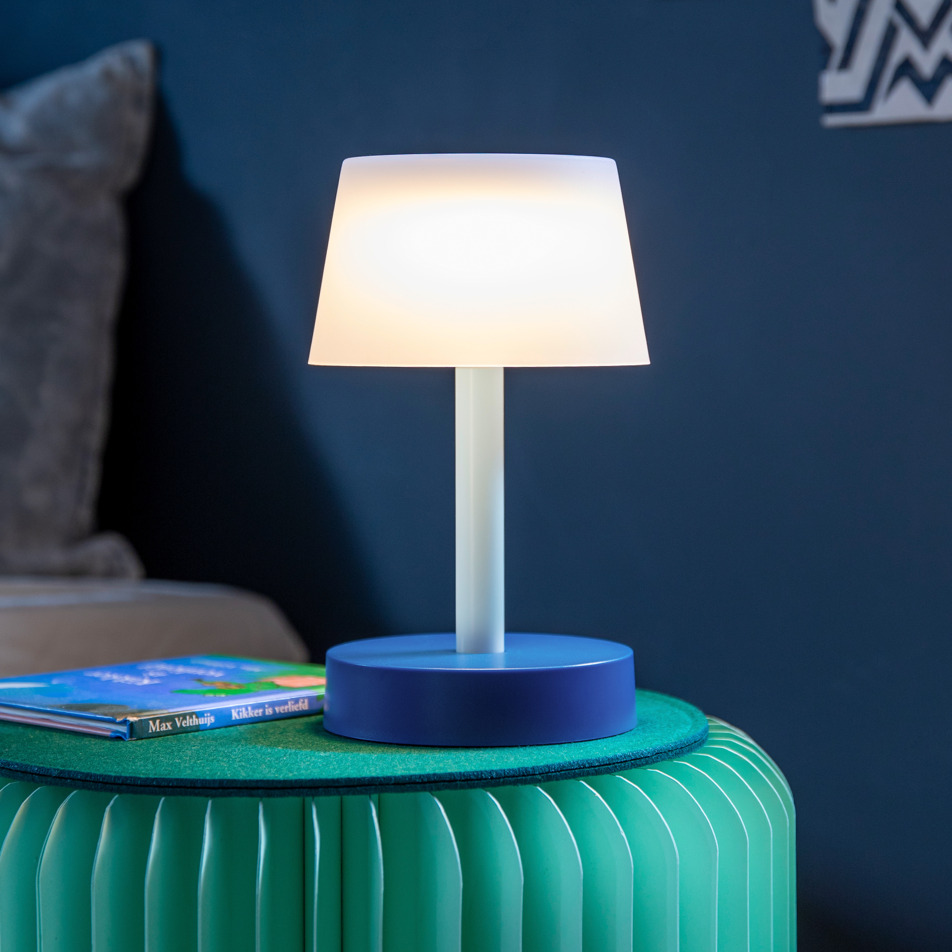 Das Produkt – eine blaue Tischleuchte 'Fritz sky' – ist auf einem grünen Hocker platziert. Die Lampe verfügt über einen weißen Schirm und emittiert warmes Licht. Ein Kinderbuch liegt daneben.
