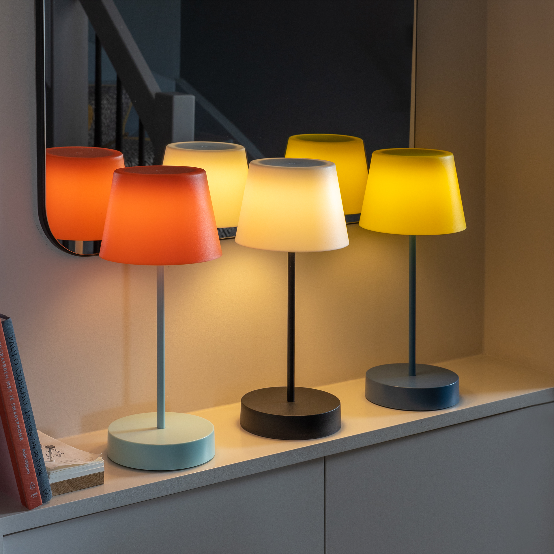 Drei eingeschaltete Tischlampen mit farbigen Lampenschirmen (orange, weiß, gelb) stehen auf einem Regal. Ein Spiegel reflektiert die Lampen. Bücher liegen links daneben.