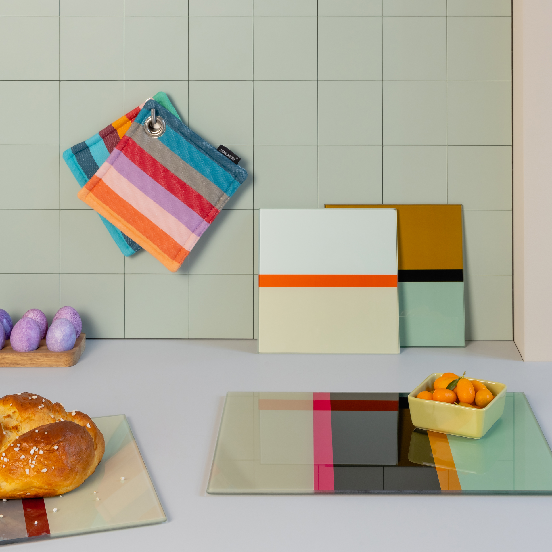 Quadratische Küchenglasplatten mit farbigen Akzenten. Im Hintergrund gestreifte Topflappen, Osterbrot und Kumquats in einer Schale.
