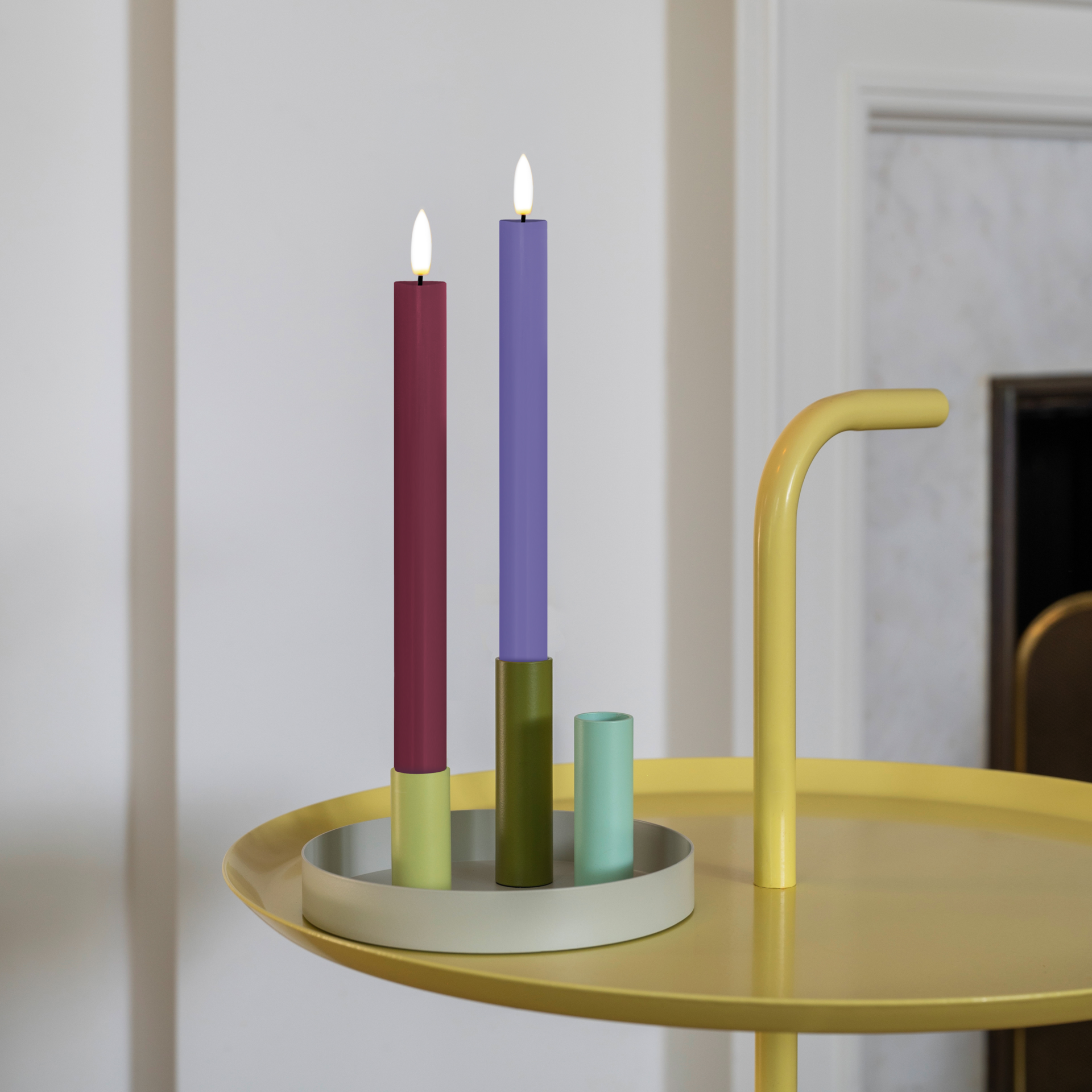 Deux bougies cylindriques à LED (rouge bordeaux et couleur lavande) sur fond blanc, avec une mèche blanche en forme de flamme et un socle blanc strié. À côté, une télécommande blanche dotée de boutons. Hauteur : 24,5 cm.