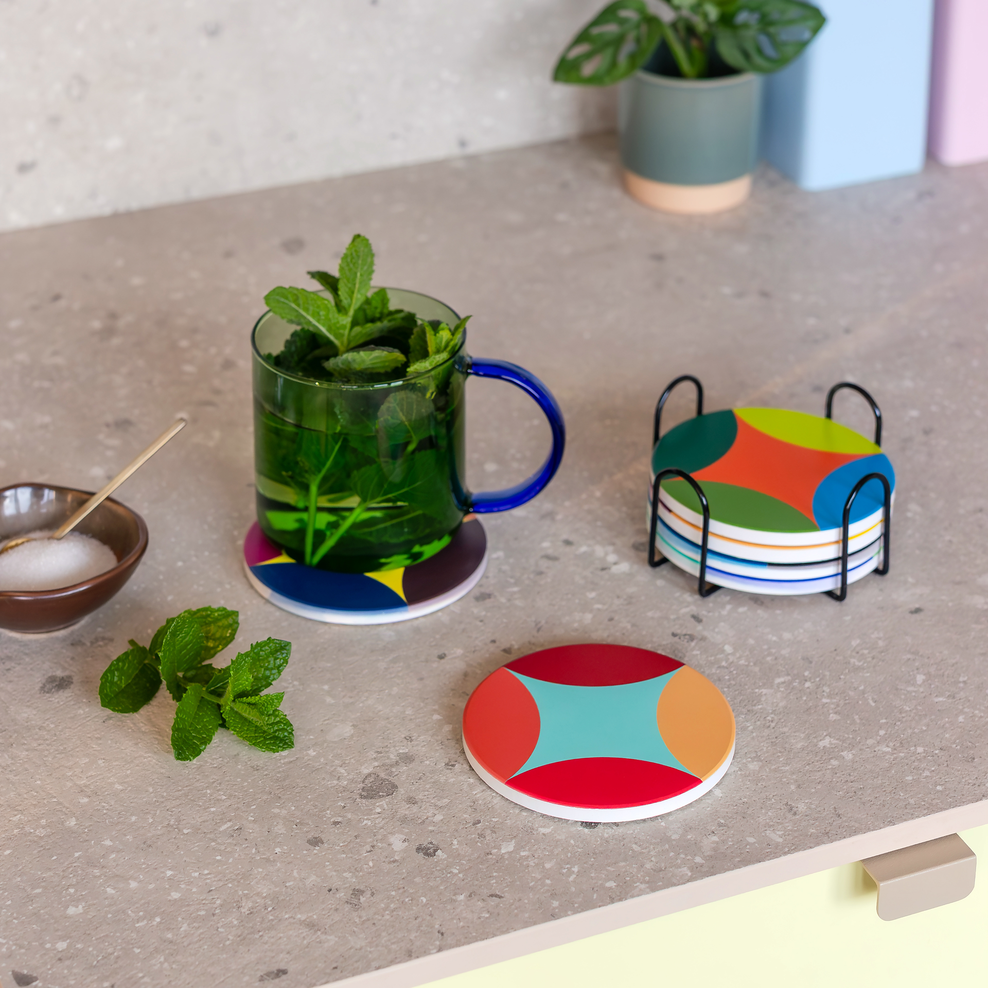 Six dessous-de-verre ronds en céramique colorée, empilés dans un support métallique noir. Chacun a un motif unique, visible sur le côté et le dessus. Dessus : bleu, orange, vert clair et vert foncé. Dessous en liège.