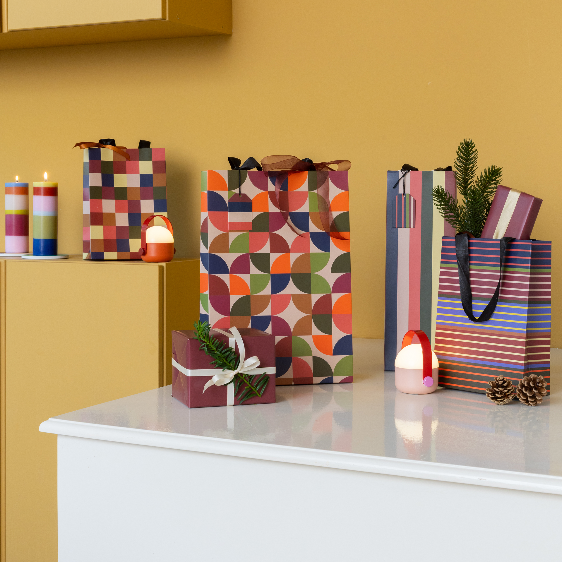 Diversi sacchetti regalo colorati con motivi vari sono disposti su una superficie bianca. Uno dei sacchetti contiene un rametto di abete. La decorazione festiva comprende anche regali incartati, candele accese e piccole lucine su uno sfondo giallo.