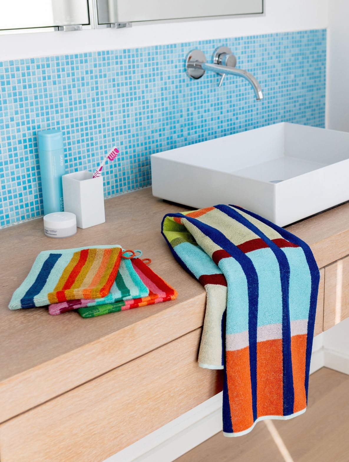 Modernes Badezimmer mit heller Holzwaschtischplatte und weißem, rechteckigem Waschbecken. Wand mit hellblauen Mosaikfliesen. Auf der Ablage bunte, gestreifte Handtücher und Waschlappen sowie blaue Flasche, Zahnputzbecher mit Bürste und kleiner Tiegel.