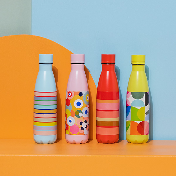 Vier farbenfrohe Trinkflaschen stehen auf einer orangefarbenen Oberfläche vor einem hellblauen Hintergrund. Jede Flasche hat ein einzigartiges Muster: Streifen, Kreise und Blumen, Linien und geometrische Formen.