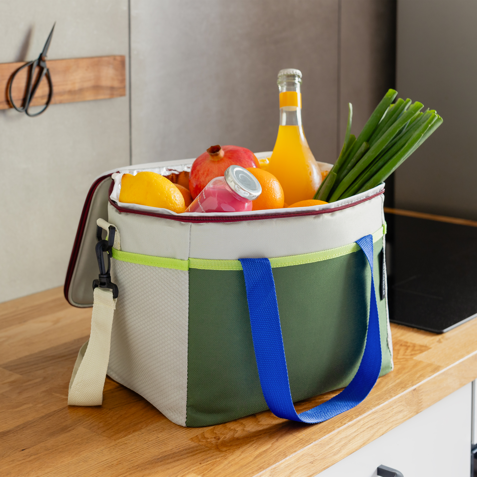 Offene Kühltasche 'Bruno' mit Inhalt: Getränke, Obst, Gemüse. Olivgrün und Beige mit blauen Trägern. Im Hintergrund Küchenzeile.