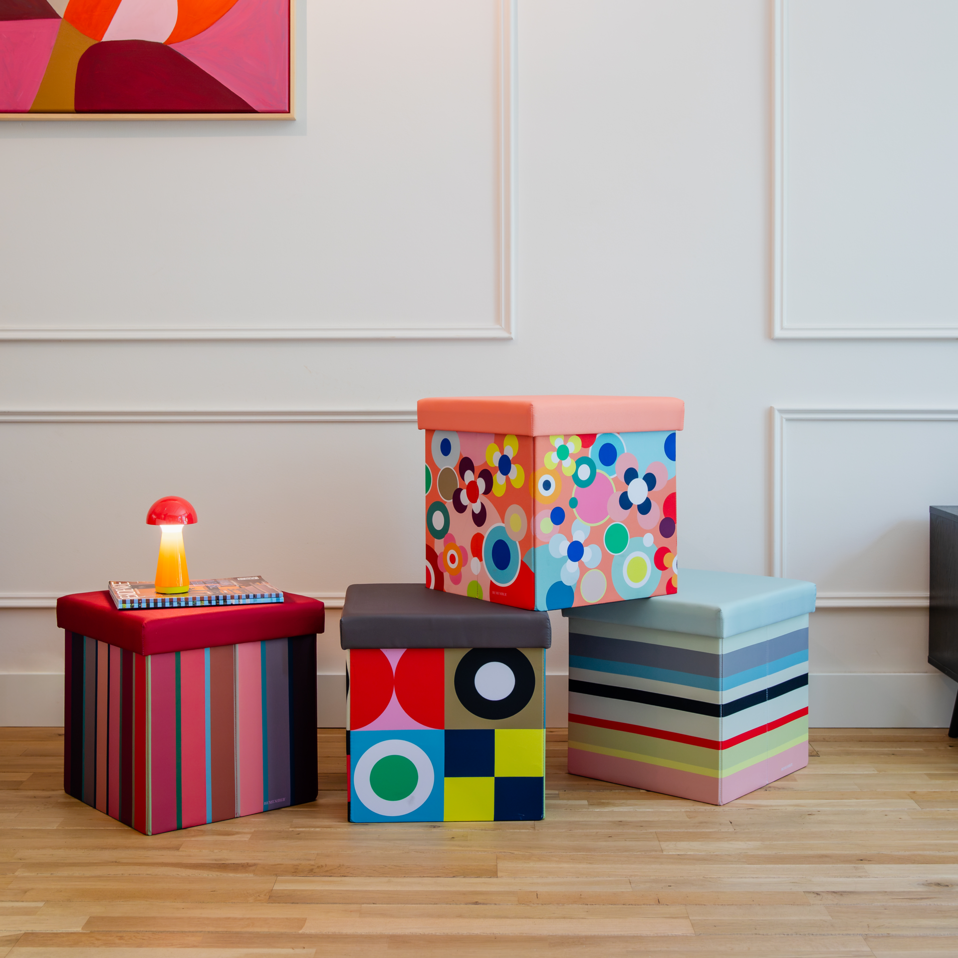 Quatre bancs colorés aux motifs variés sur un parquet clair devant un mur blanc. À gauche, un banc à rayures rouges avec lampe champignon et magazines. Au centre, motif géométrique et floral. À droite, banc à rayures. REMEMBER®, rangements.