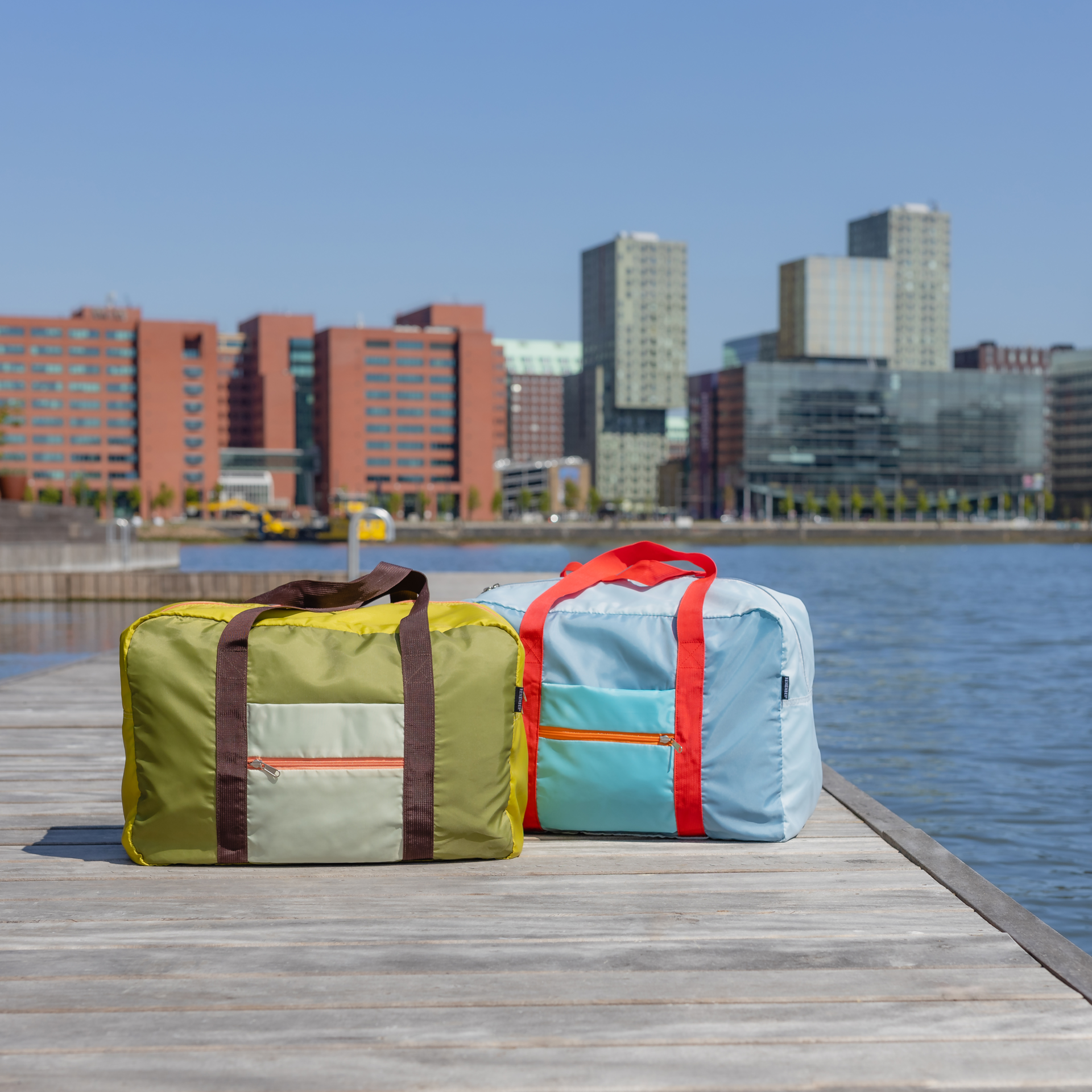 Rechteckige Reisetasche 'Tom' in Hellblau und Grau mit roten Tragegriffen. Eine aufgesetzte, hellblaue Tasche mit orangefarbenem Reißverschluss befindet sich auf der Vorderseite.