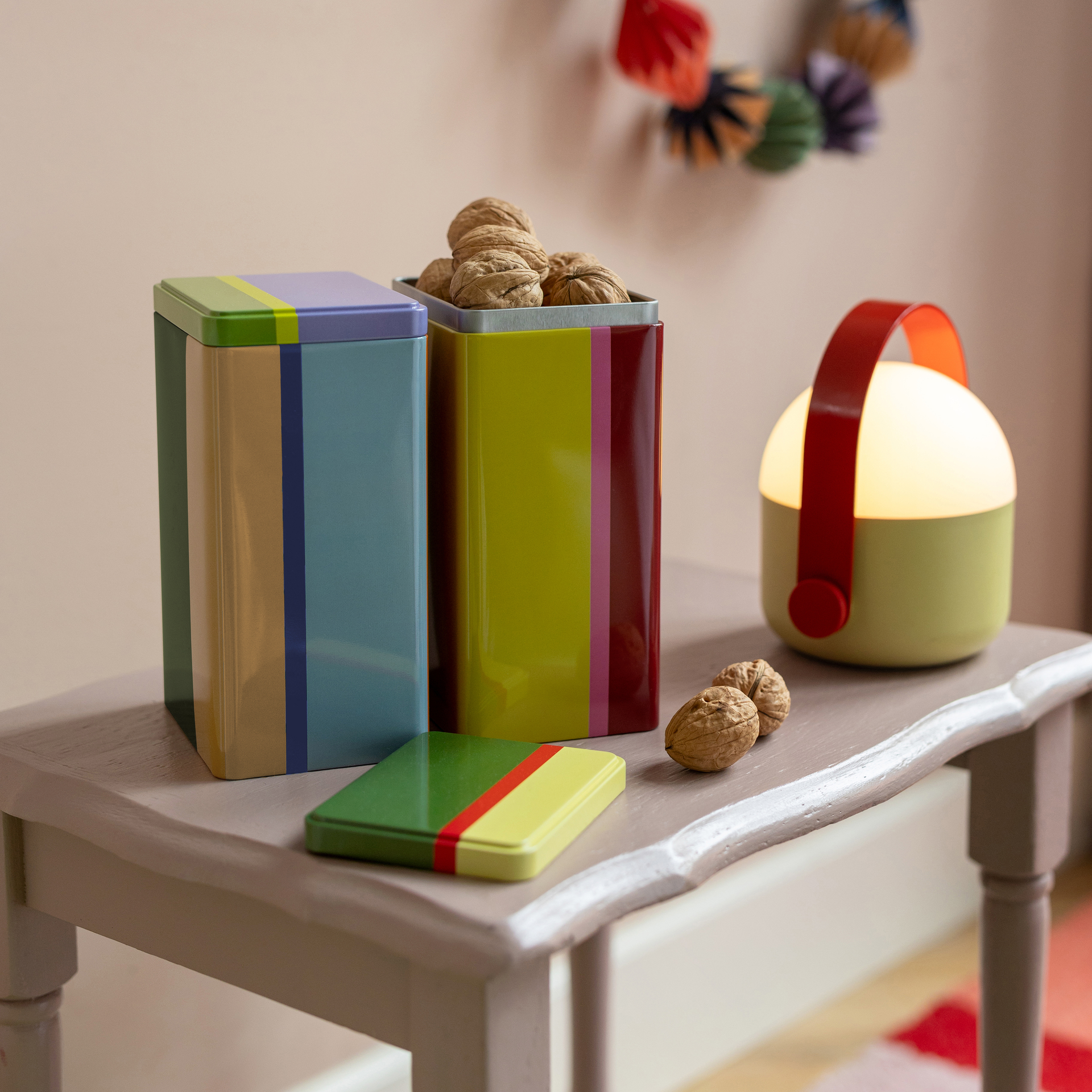 Set di lattine colorate "Yara" su tavolo con noci e lampada. Le lattine sono a strisce blu, gialle, verdi e rosse.
