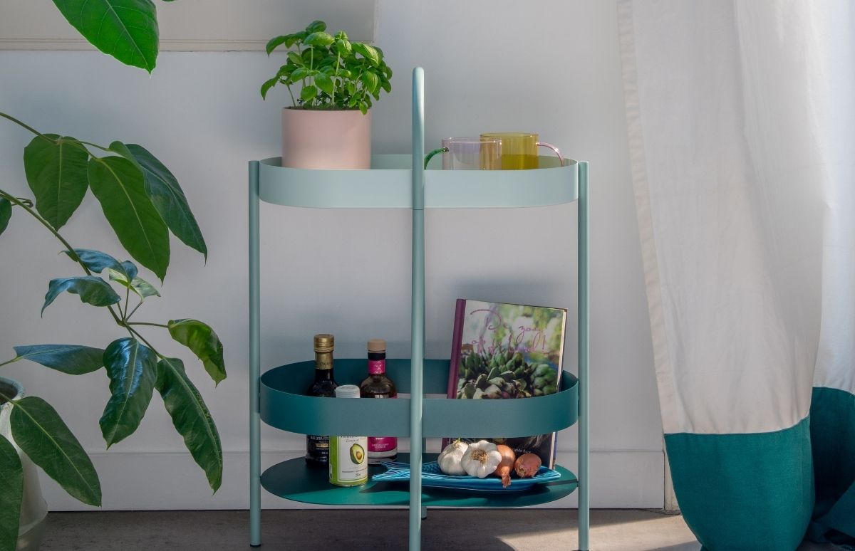 Hellblauer Servierwagen mit zwei Ebenen. Oben ein Basilikumtopf und zwei bunte Tassen. Unten Kochzutaten: Öle, Avocadoöl-Dose, Kochbuch, Knoblauch und Zwiebeln auf blauer Schale. Im Hintergrund Pflanze und Vorhang.