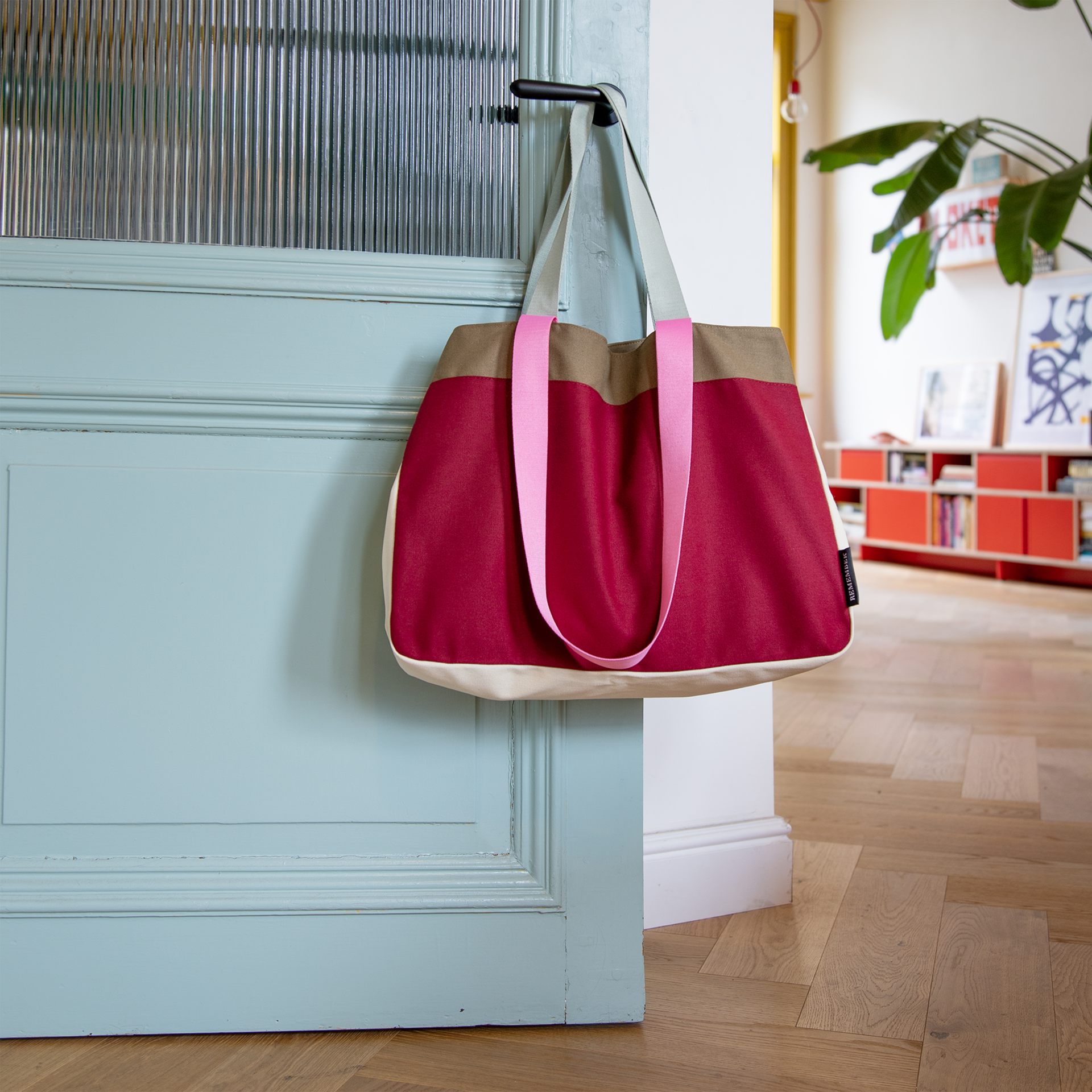 Canvas Tasche 'Emma' in Rot, Beige und Khaki hängt an einer hellblauen Tür. Die Henkel sind rosa und hellgrau. Im Hintergrund sind ein Holzboden und ein Regal sichtbar.