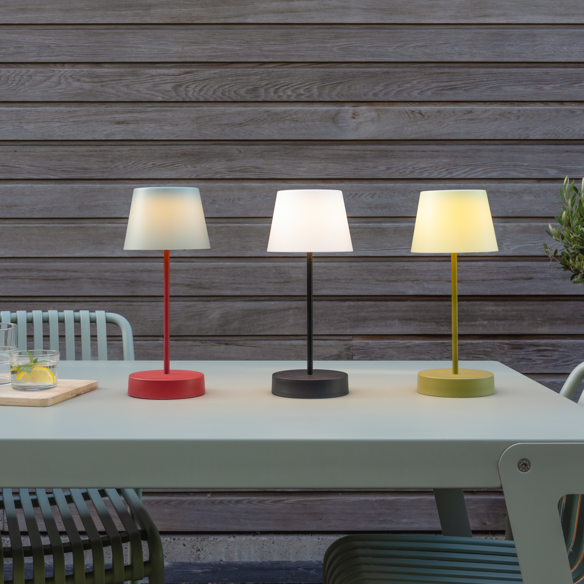 Drei Tischleuchten 'Oscar' in rot, schwarz und gelb auf einem Gartentisch. Jede Lampe leuchtet in einer anderen Helligkeitsstufe. Im Hintergrund ist eine Holzwand zu sehen.