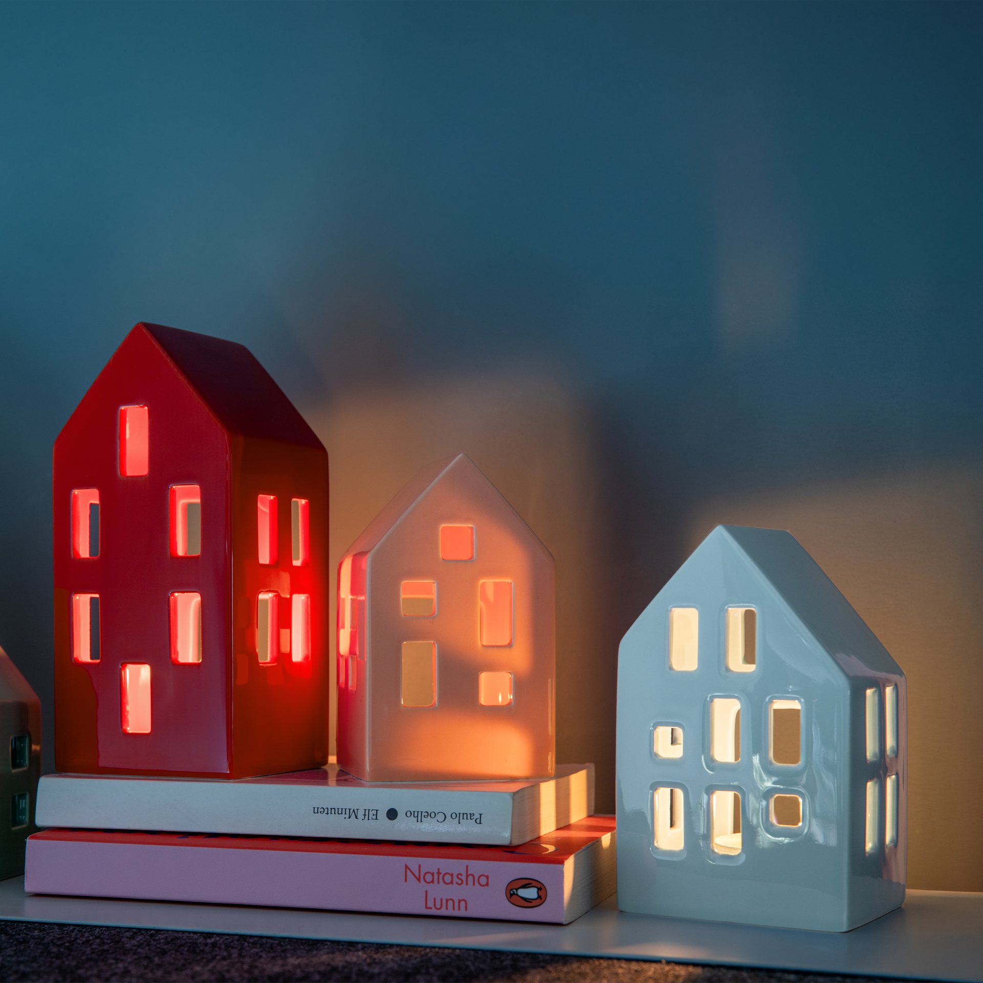 Mehrere Teelichthäuser Nyhavn in rot, peach und blau stehen auf Büchern. Durch die Fenster scheint warmes Licht.