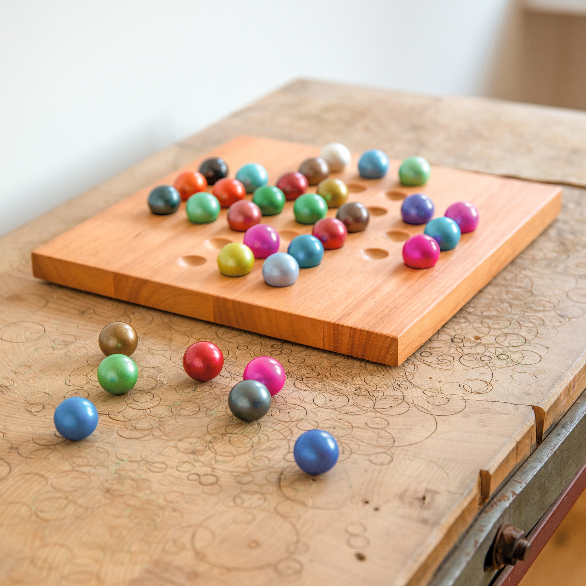 Planche en bois avec des trous, remplie de billes de verre colorées. Quelques billes sont posées à côté. Le jeu de solitaire est posé sur une table avec des gravures.