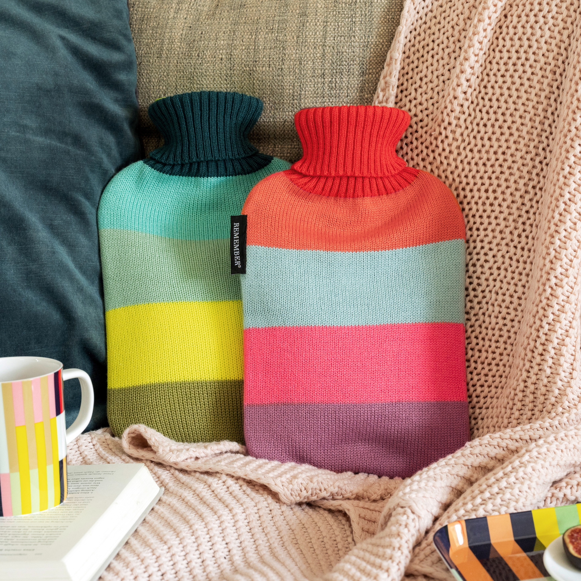 Zwei bunte 'Pepe' Wärmflaschen mit gestreiften Strickbezügen liegen auf Sofa und Decke. Eine Tasse und Buch sind im Bild.
