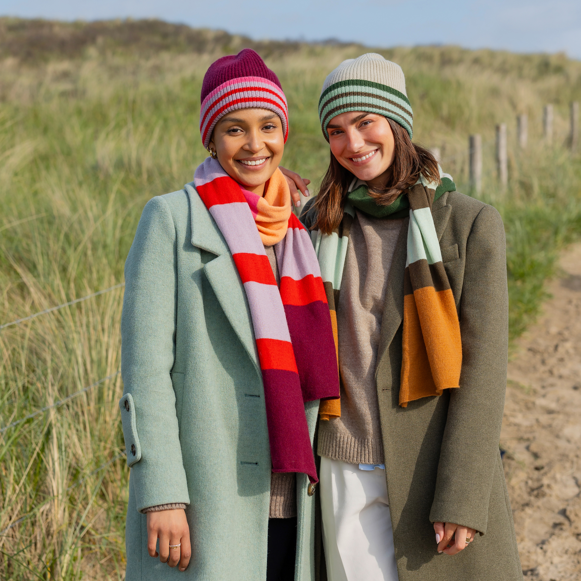 Zwei lächelnde Frauen mit Woll-Kaschmir-Mützen und farblich passenden Schals stehen vor einer Dünenlandschaft.
