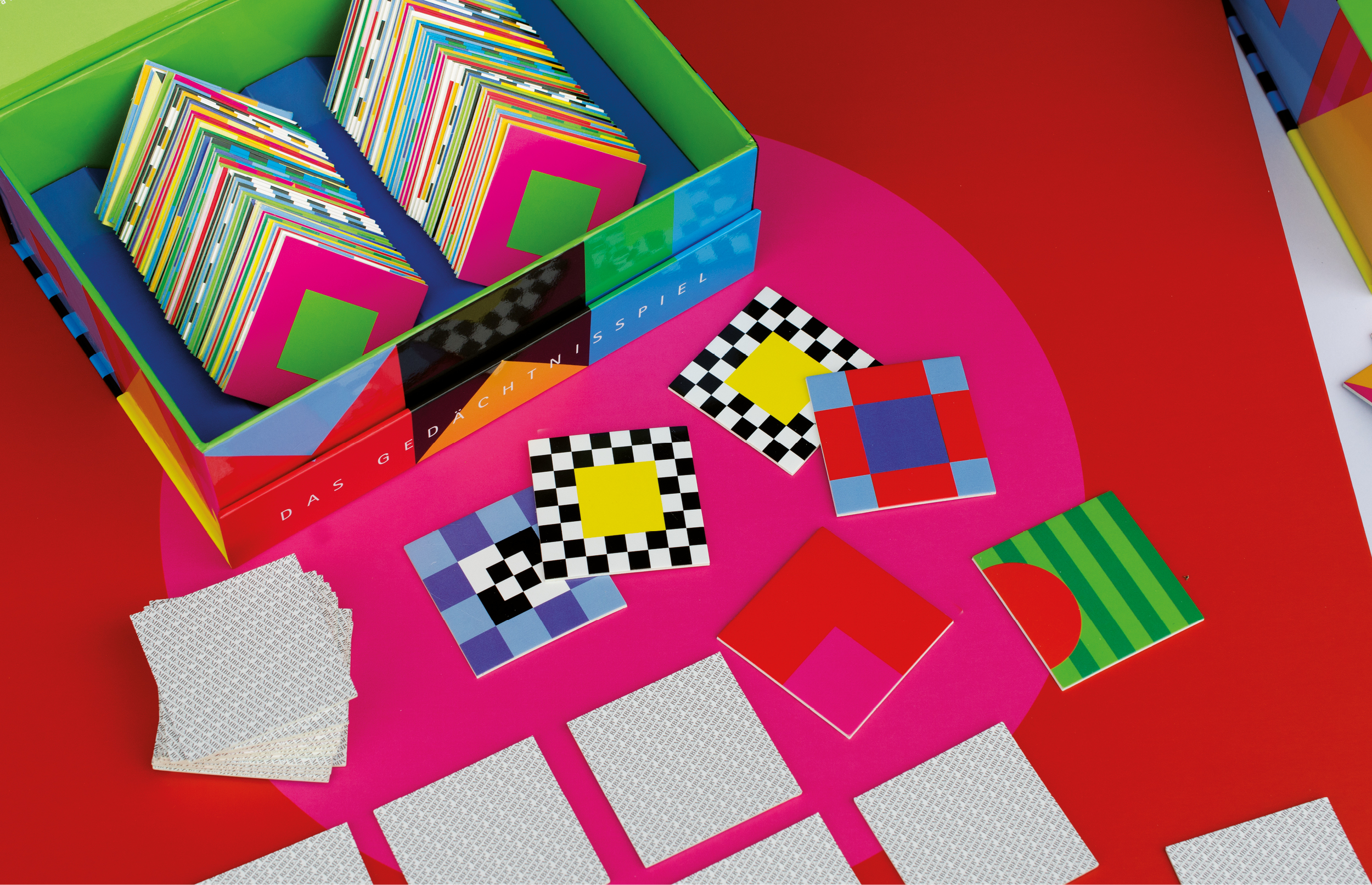 Spielkarten und eine bunte Schachtel eines Gedächtnisspiels liegen auf einem roten und pinkfarbenen Hintergrund. Die Karten zeigen bunte geometrische Muster.
