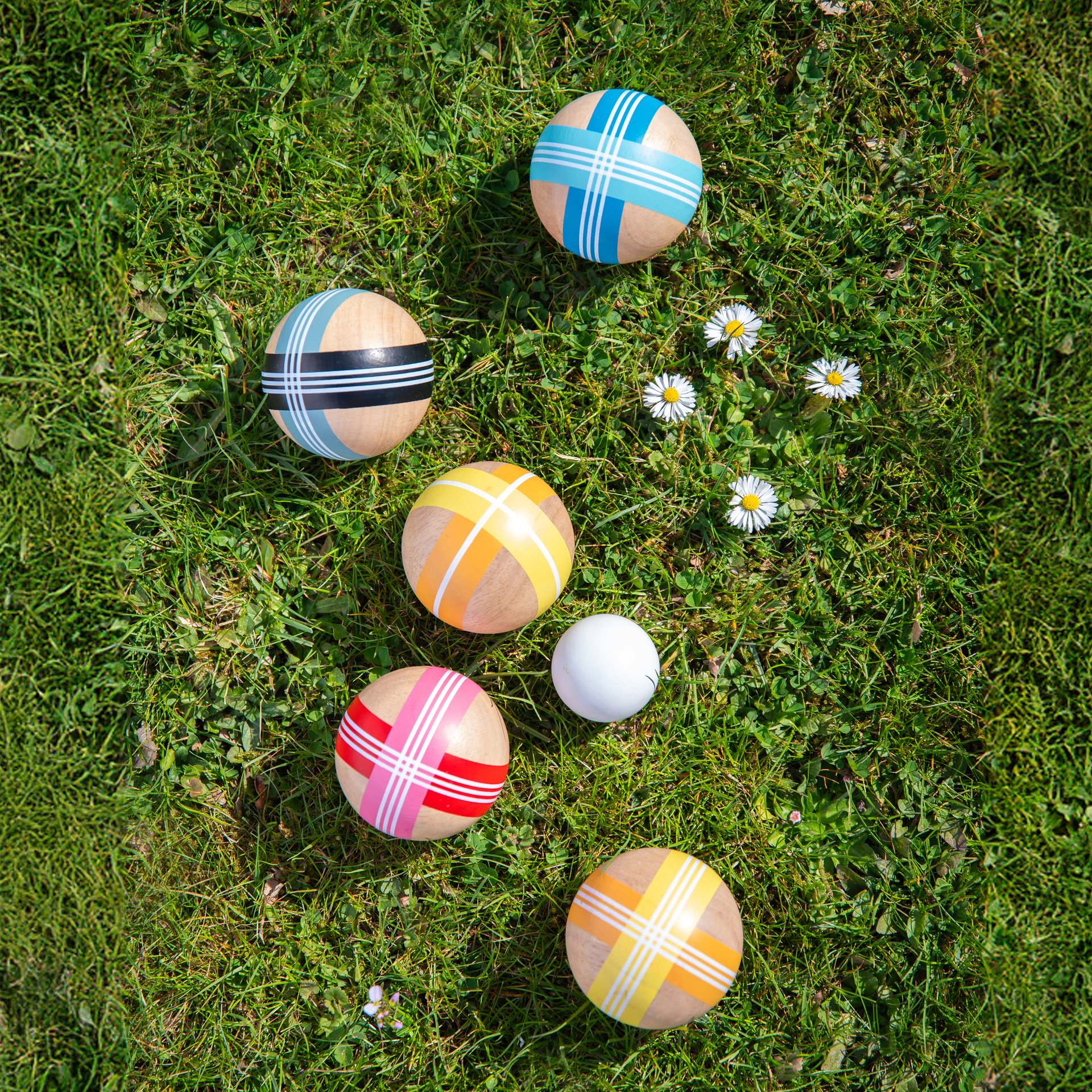 Draufsicht: Boccia-Kugeln mit Streifenmuster und Zielkugel liegen auf Gras. Einige Gänseblümchen sind zwischen den Kugeln verstreut.