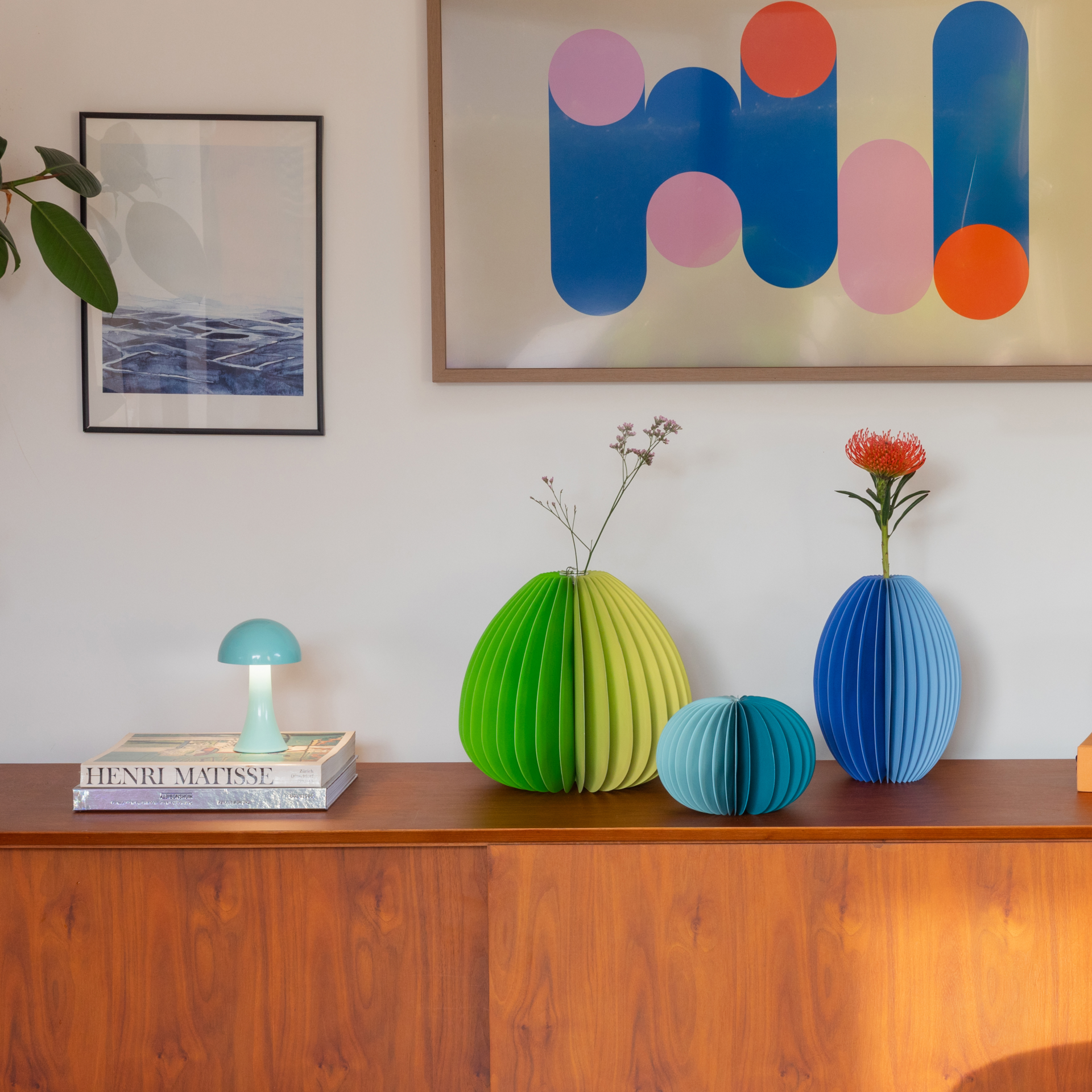 Holzkommode mit farbigen Vasen und Kunstwerken. Eine grüne, blaue und hellblaue Vase stehen auf der Kommode. Darüber hängen zwei Bilder: ein abstraktes und ein Landschaftsbild. Eine Lampe steht links.